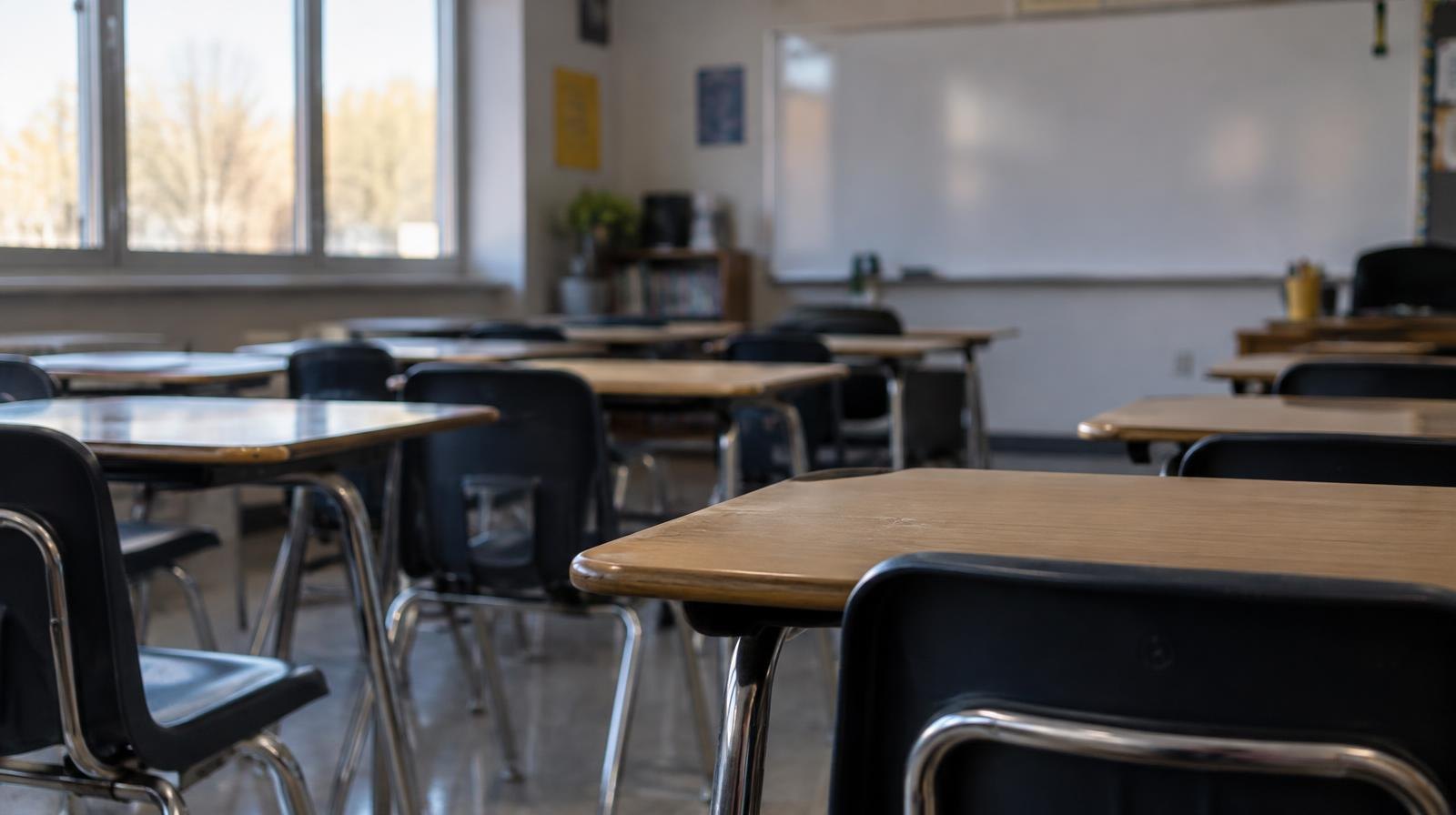 Rows of empty desks and chairs in a sunlit high school classroom with a whiteboard at the front.