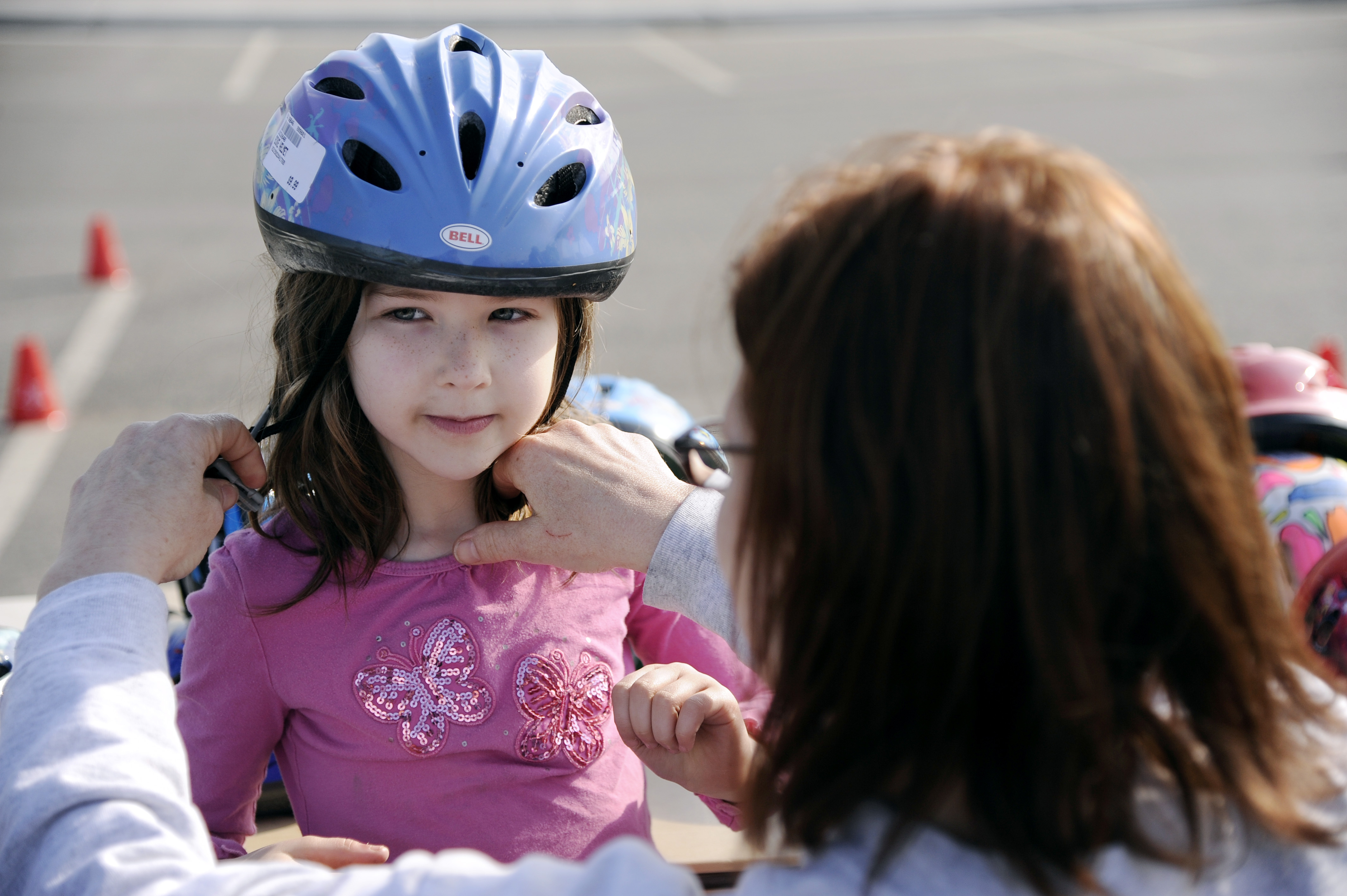 Adult helping a young girl put on a blue bicycle helmet at an outdoor event.