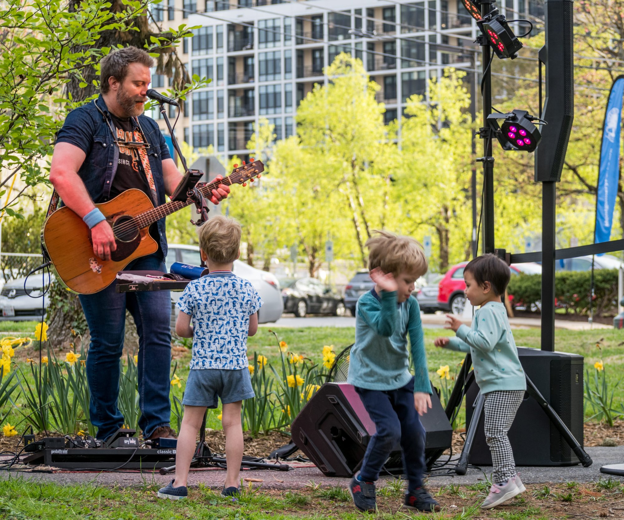 Guitarist performs on outdoor stage while three young children dance in front of him.