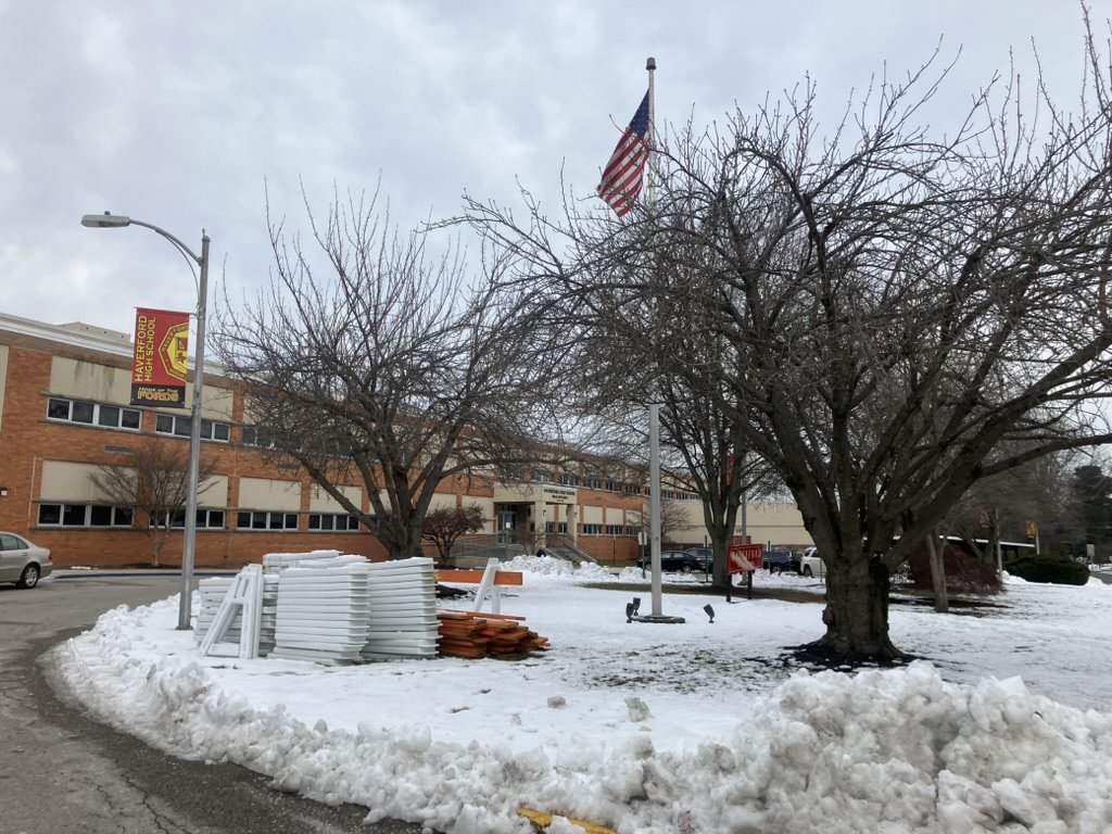 Brick school building entrance in winter with snow, a flagpole, and bare trees.
