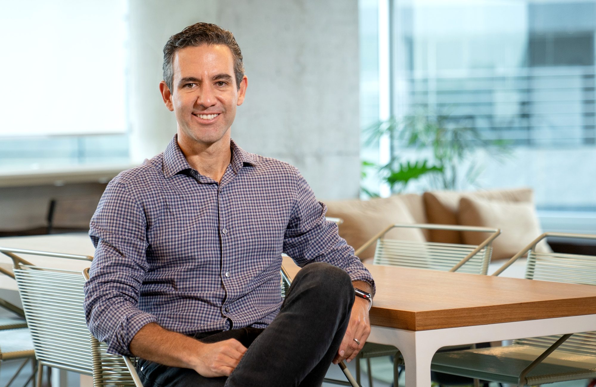A man in a checkered button-down shirt sits smiling in a modern office lounge area with a wood table and greenery in the background.