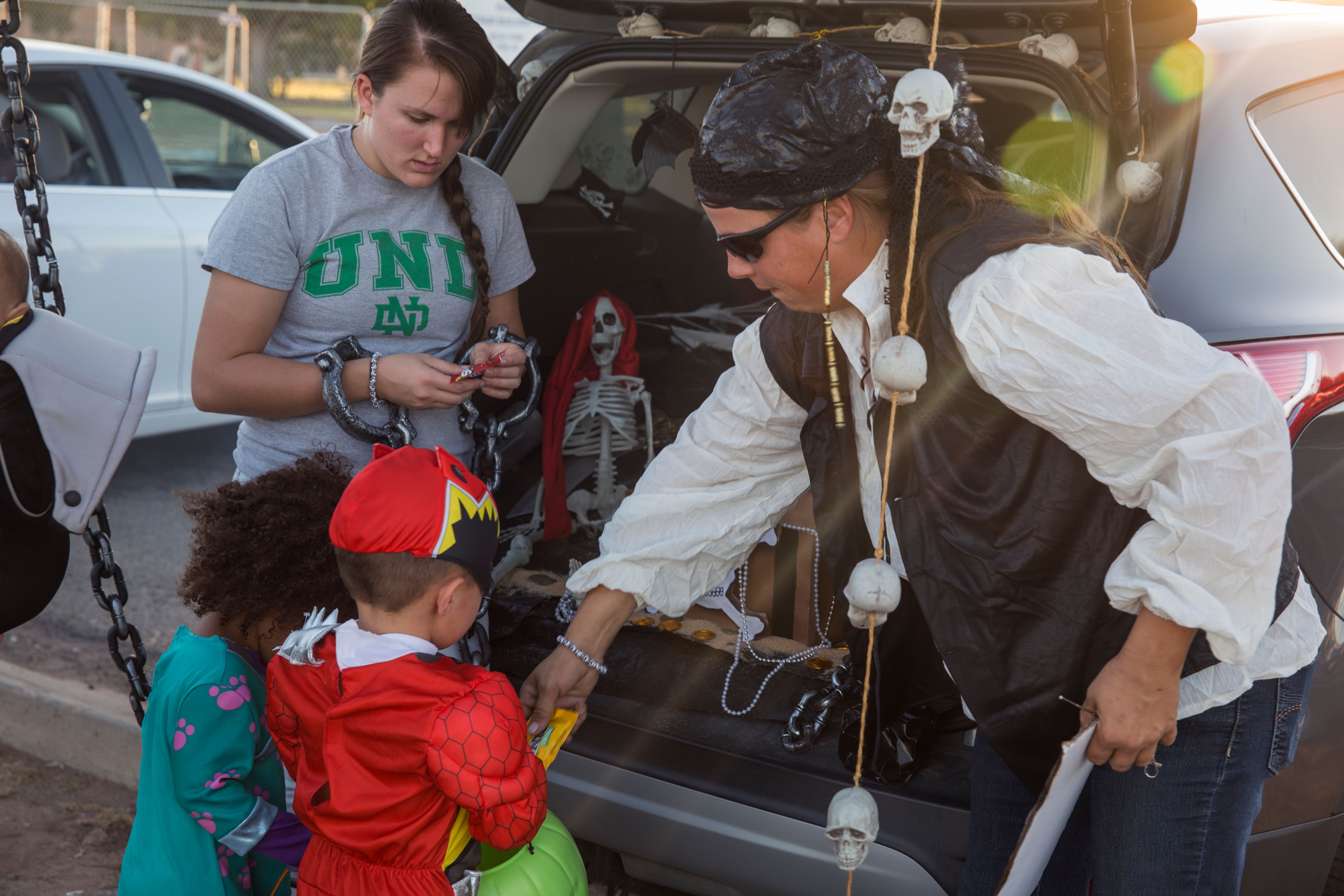 Children in Halloween costumes receive candy from a decorated car trunk outdoors.