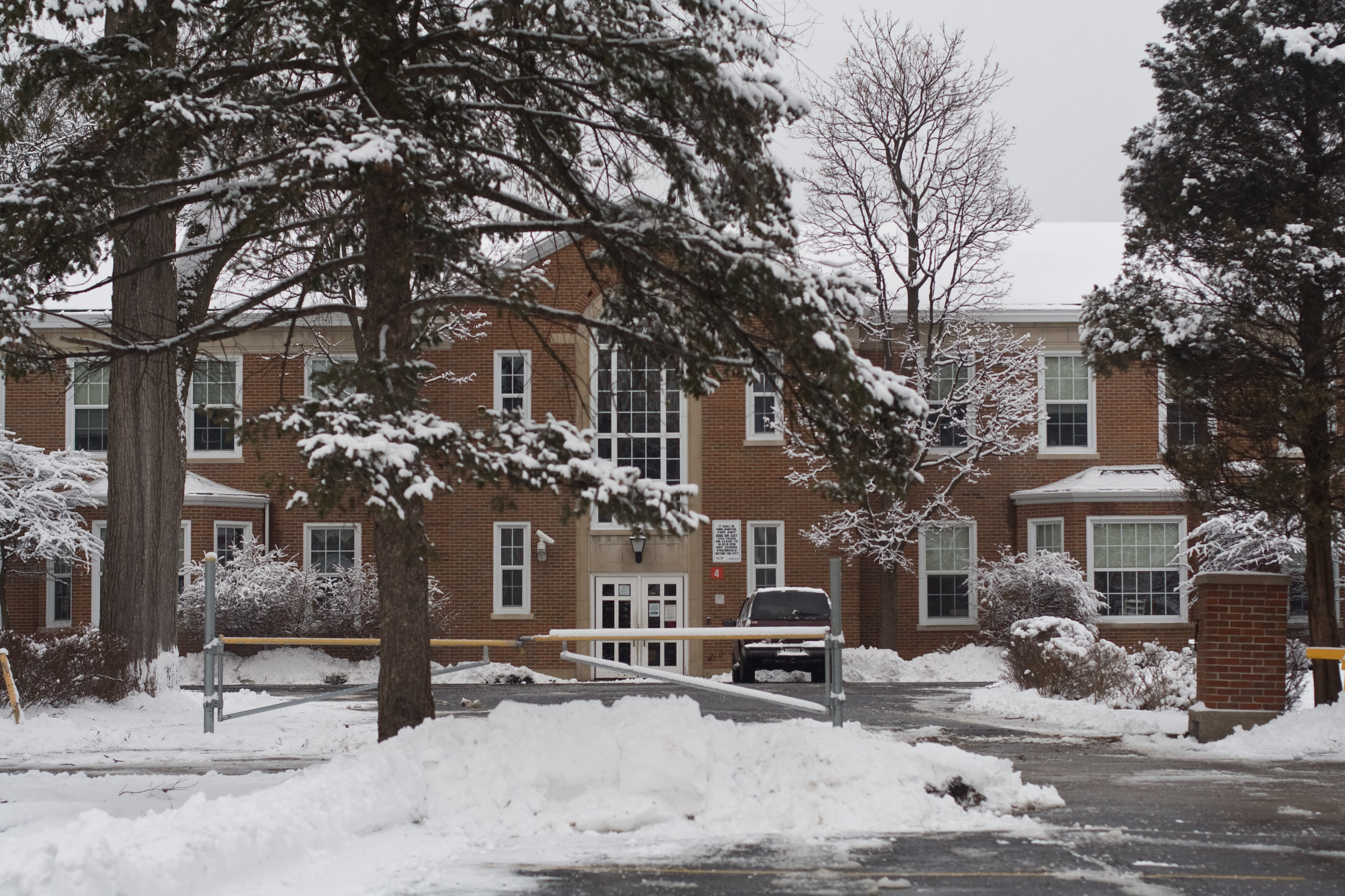 Two-story brick school building behind snow-covered evergreen trees in winter.