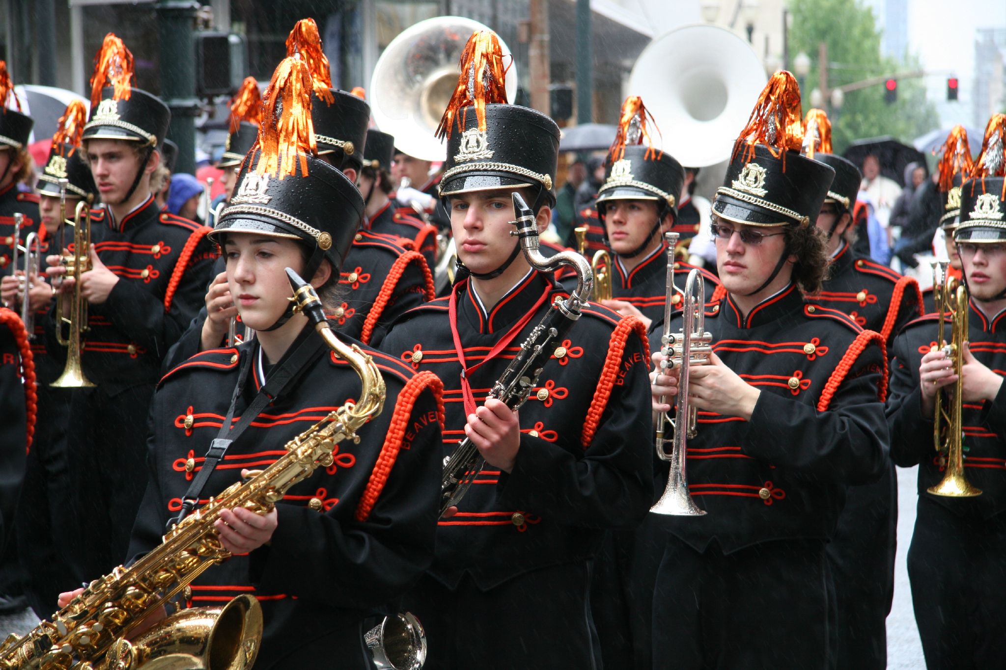 Marching band students in black and red uniforms playing brass and woodwind instruments outdoors.