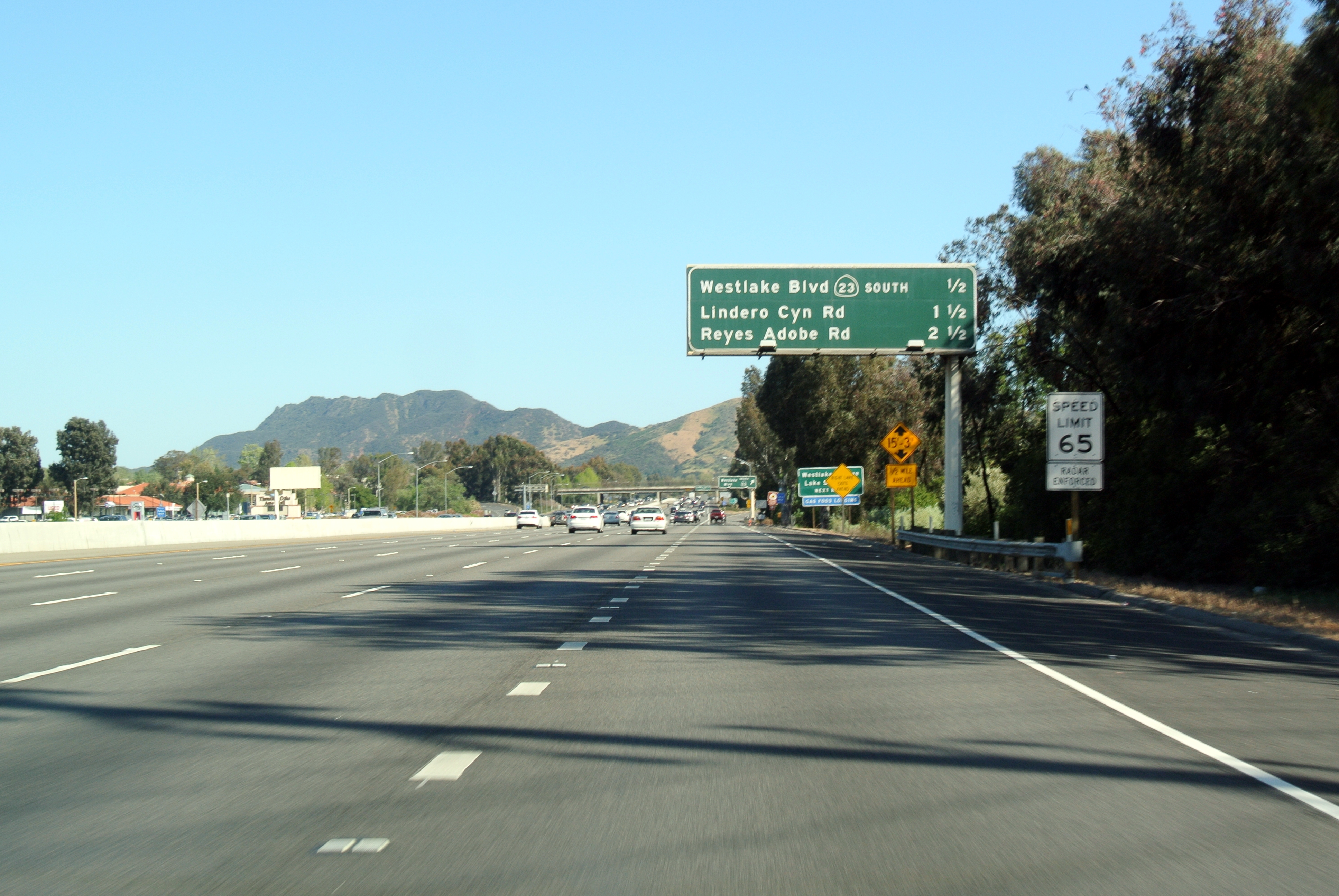 Multi-lane freeway with overhead green highway signs and mountains in the background.