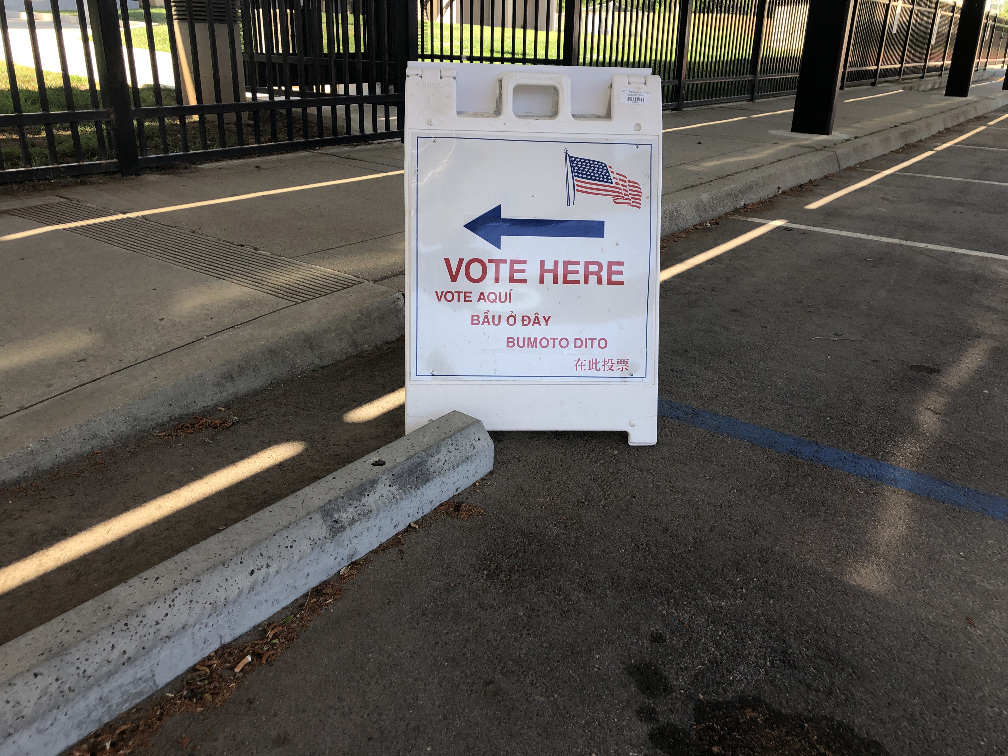 A-frame sign reading Vote Here in five languages stands in a parking lot.
