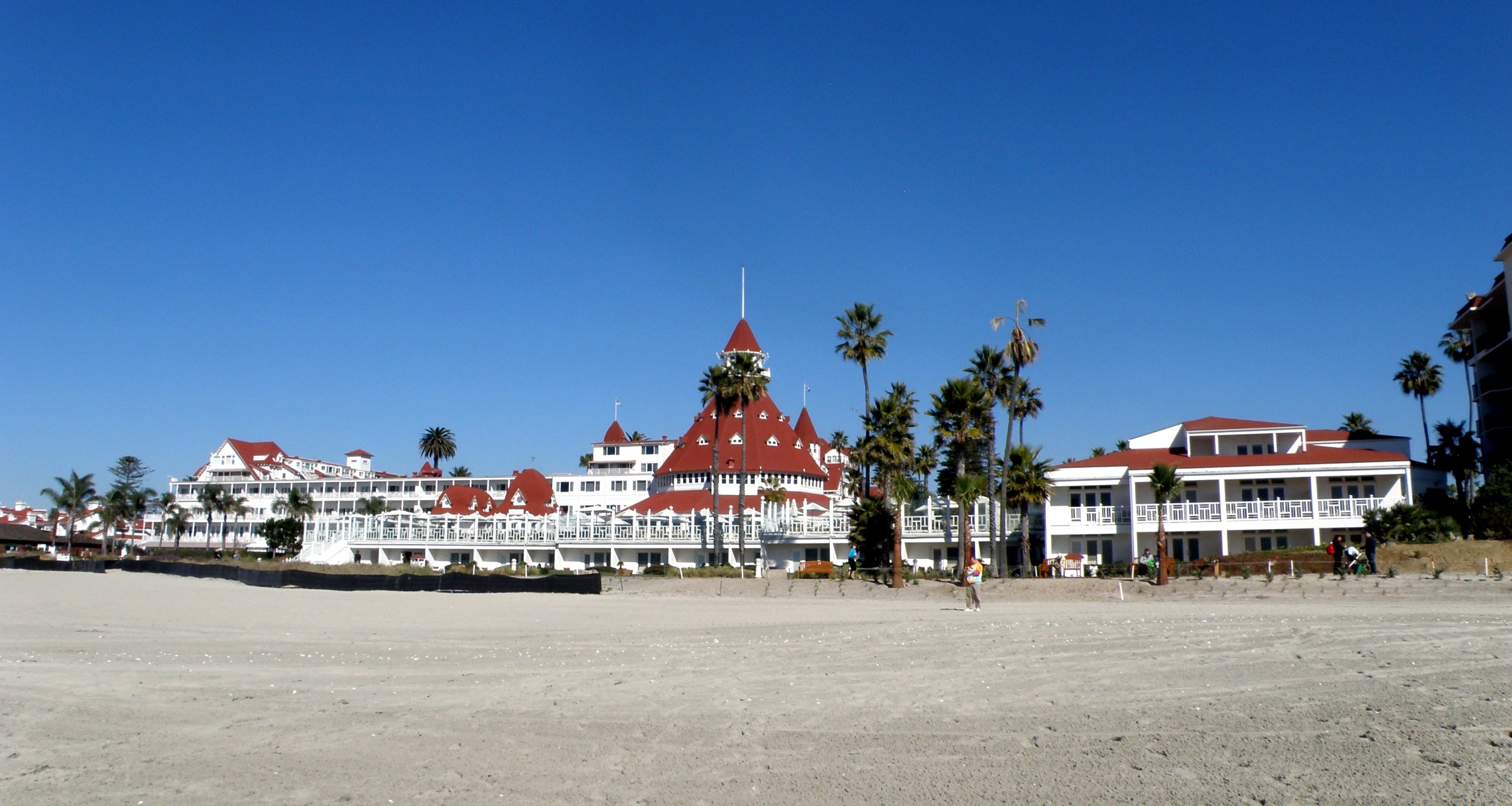 Victorian resort hotel with red roofs seen from a sandy beach with palm trees.