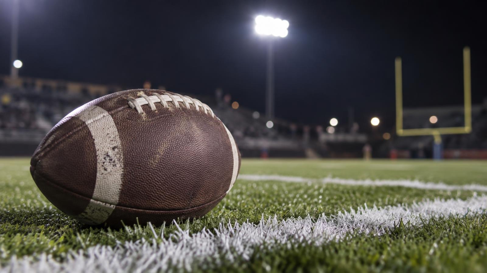 An American football on green grass near a white yard line under stadium lights.