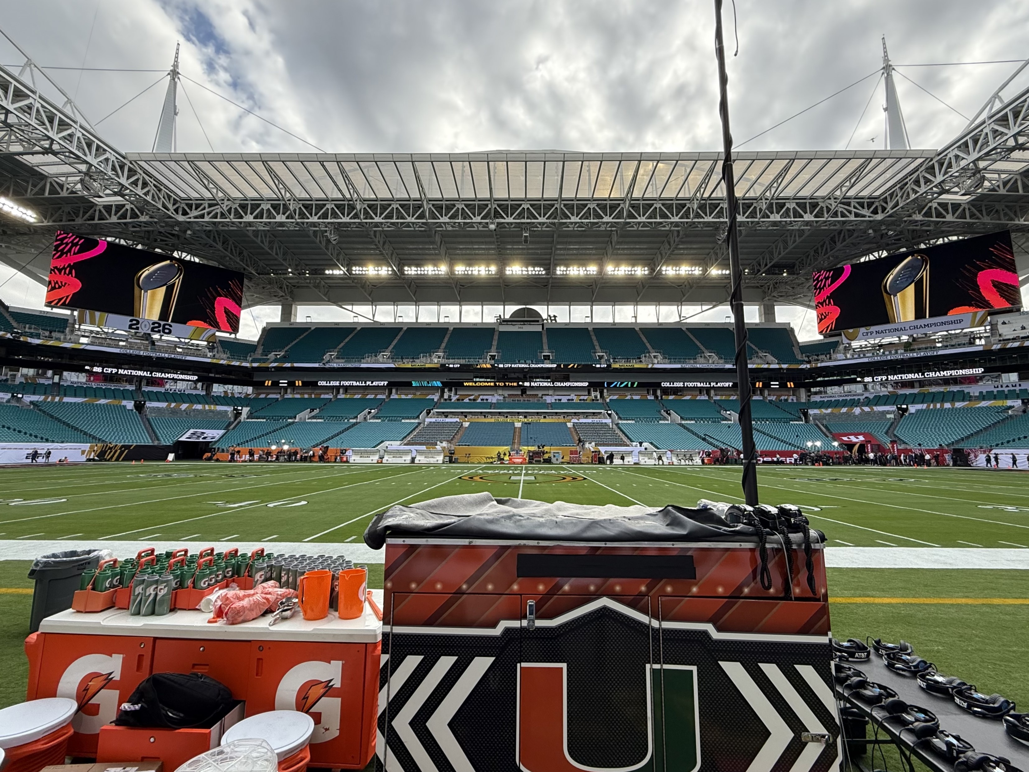 Stadium interior viewed from a football sideline with team equipment and video boards visible.