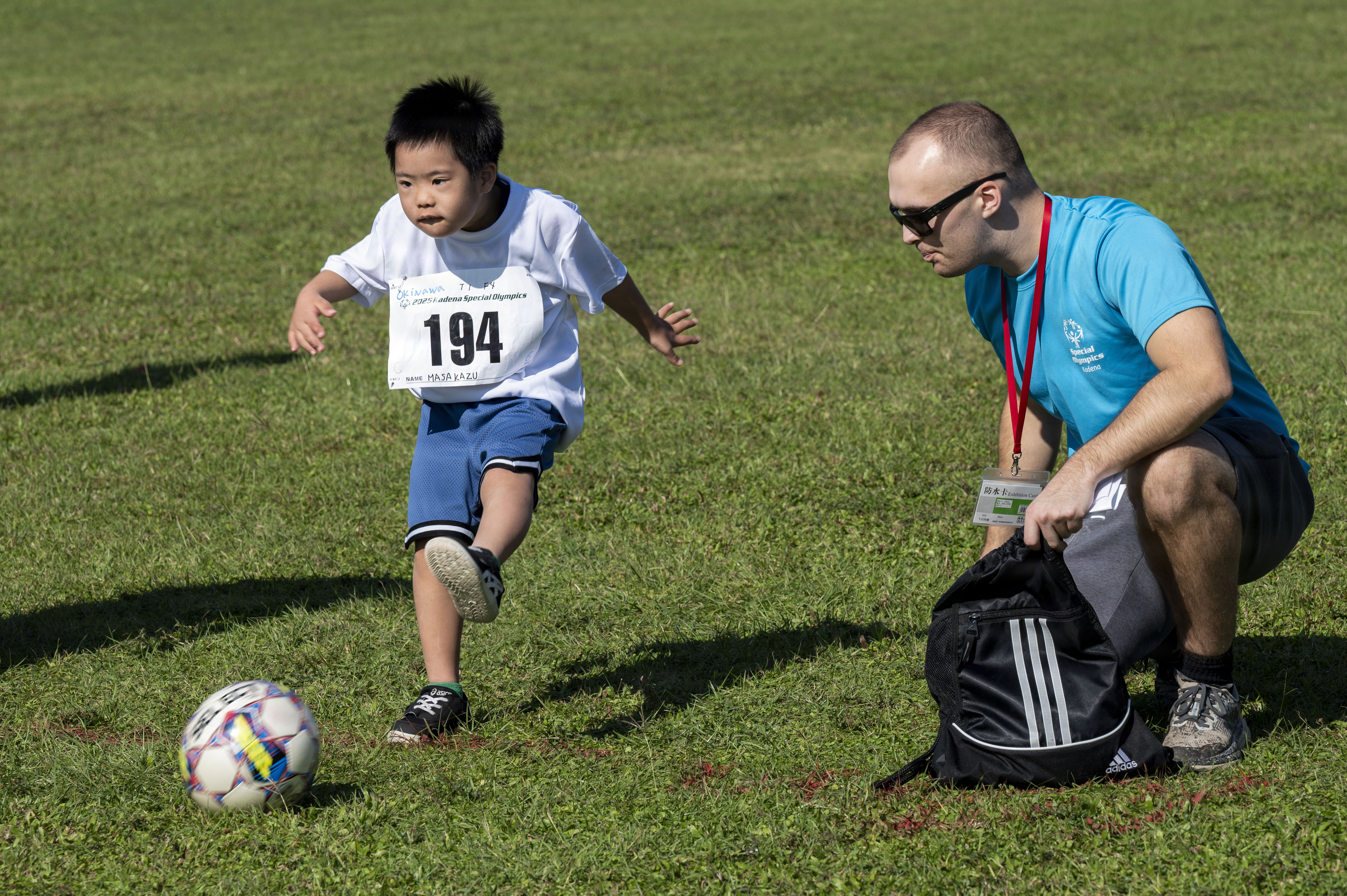 Young athlete in a numbered bib kicking a soccer ball while a coach crouches nearby on a grass field.