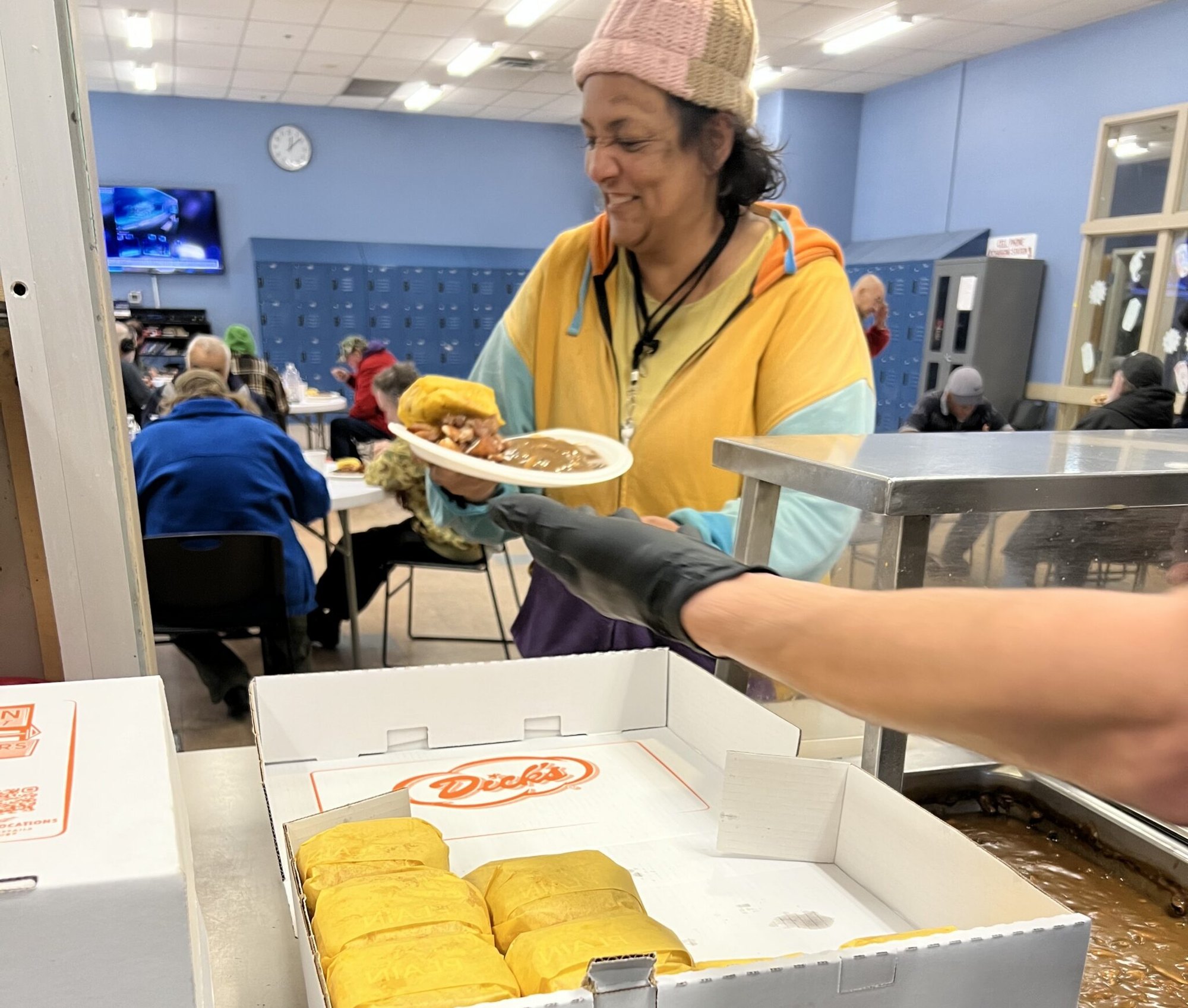 Person in yellow hoodie receiving food at a shelter serving line, Dick's burger box in foreground.