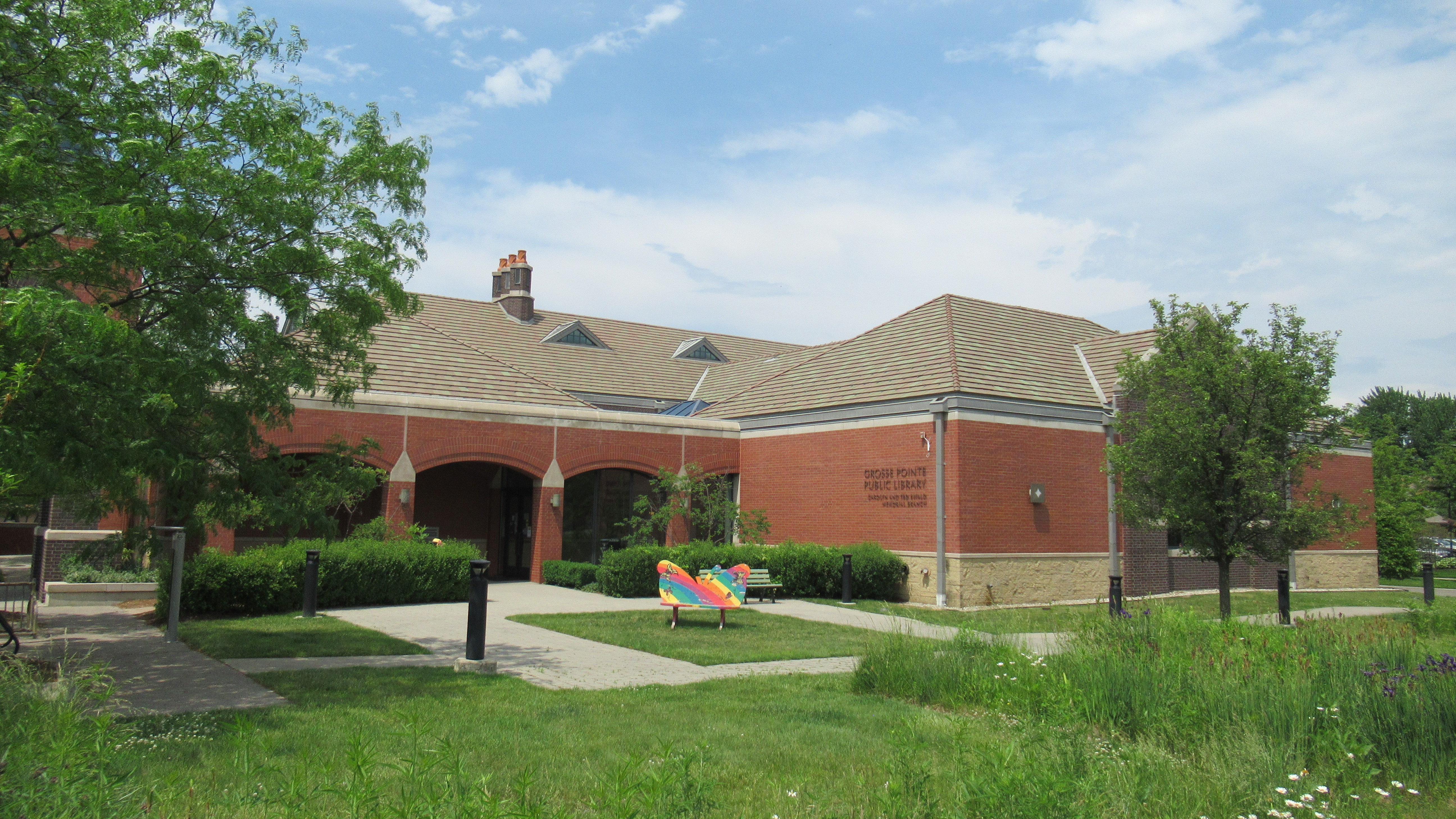 Single-story brick library building with arched entrance and sculpture on a sunny day.