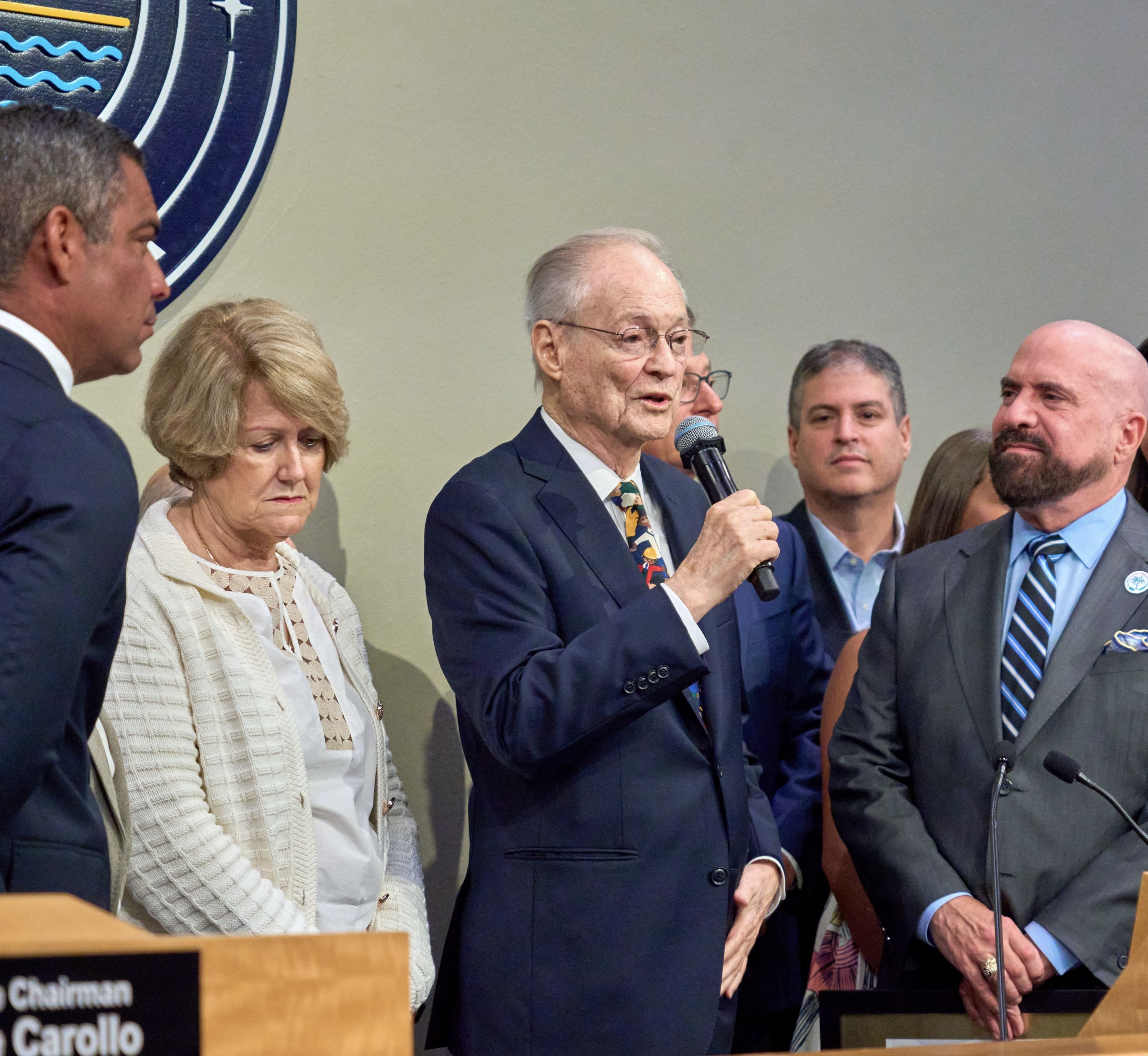 Officials at a podium inside a government commission chamber with a municipal seal on the wall.