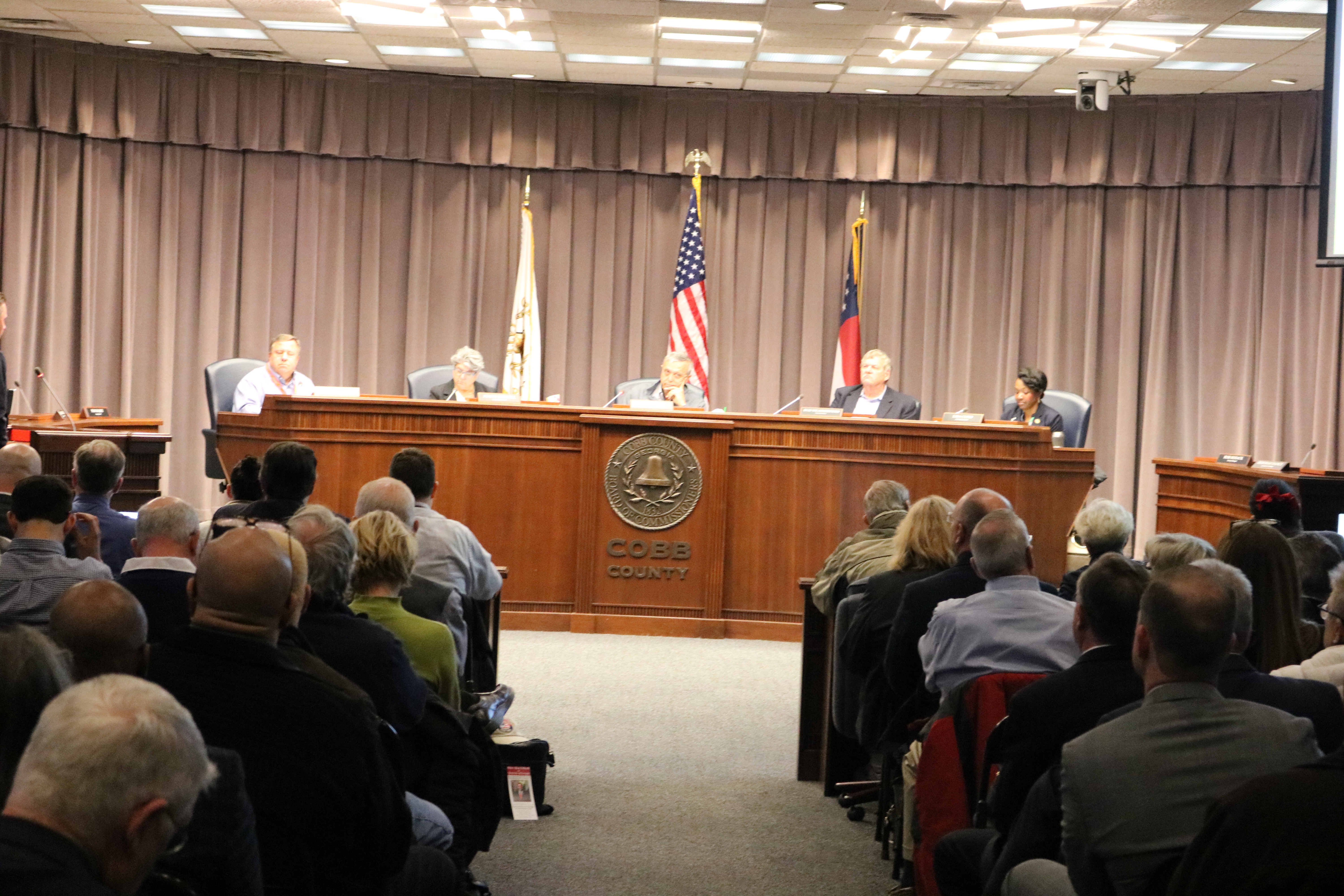 Commissioners seated at a dais with a county seal, facing a packed public gallery.