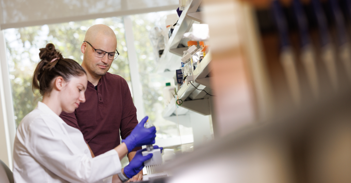 A man in glasses and a maroon shirt observes a woman in a white lab coat working with laboratory equipment.