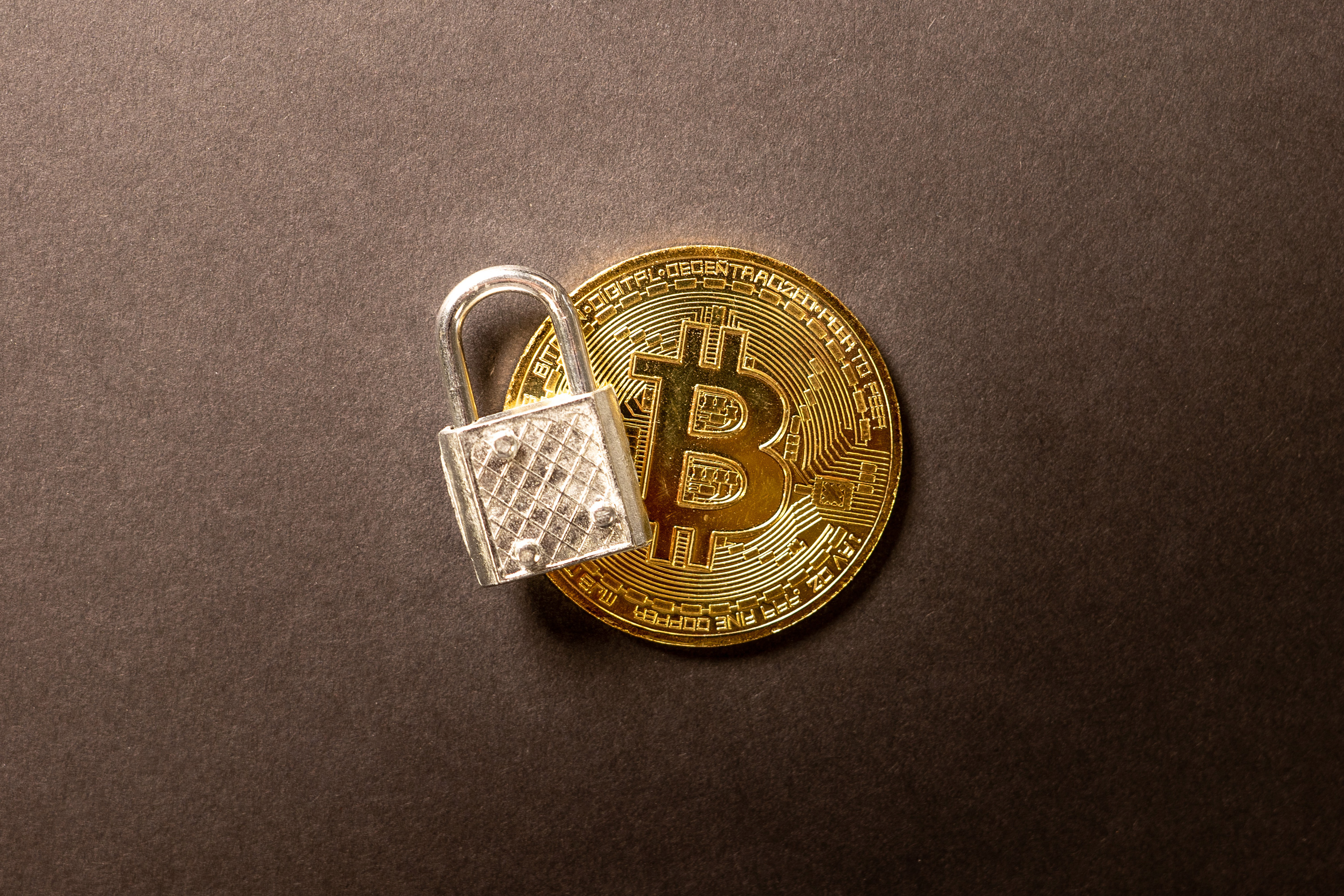 A gold Bitcoin coin with an open silver padlock leaning against it on a dark surface.