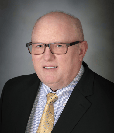 Terence Collins wears glasses, a dark jacket, a checked shirt and a yellow patterned tie against a gray background.