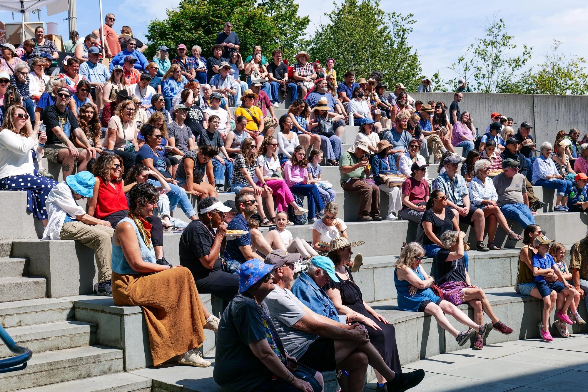 Large diverse crowd seated on tiered concrete steps outdoors on a sunny day.
