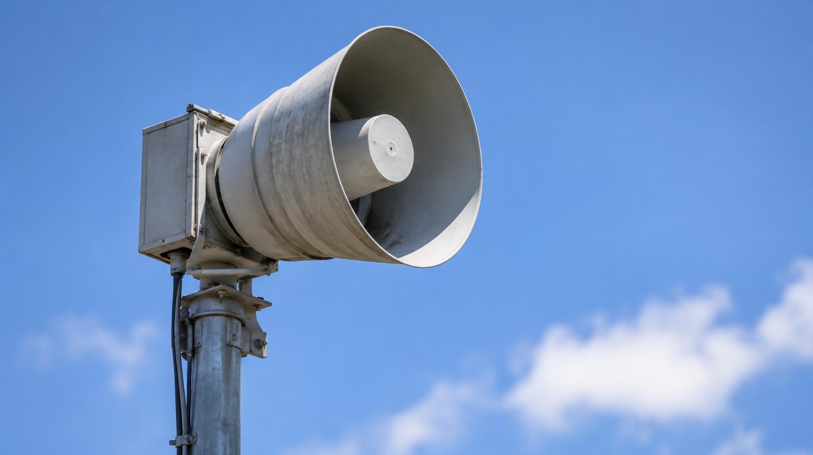 An outdoor emergency warning siren on a tall metal pole against a blue sky.