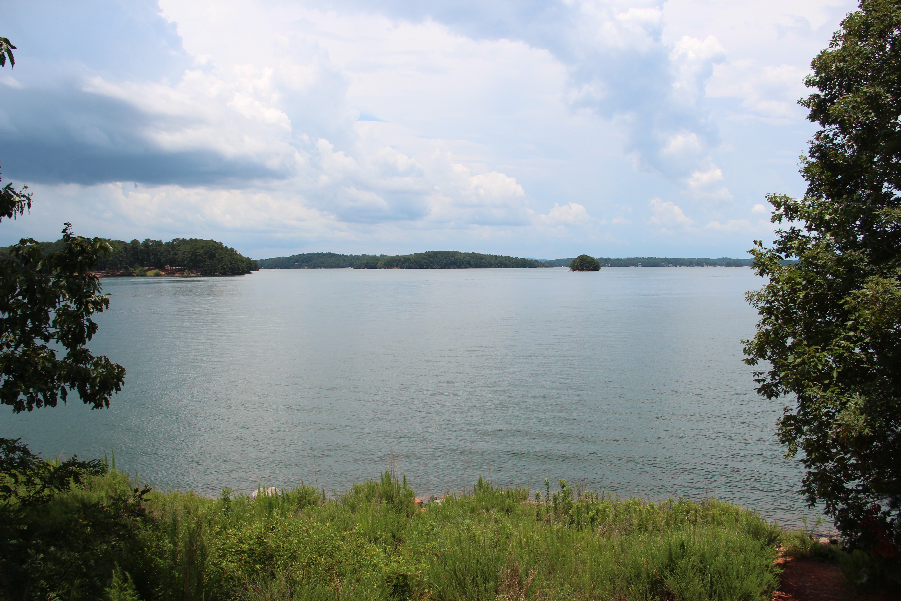 A wide, calm lake surrounded by forested shorelines under a partly cloudy summer sky.