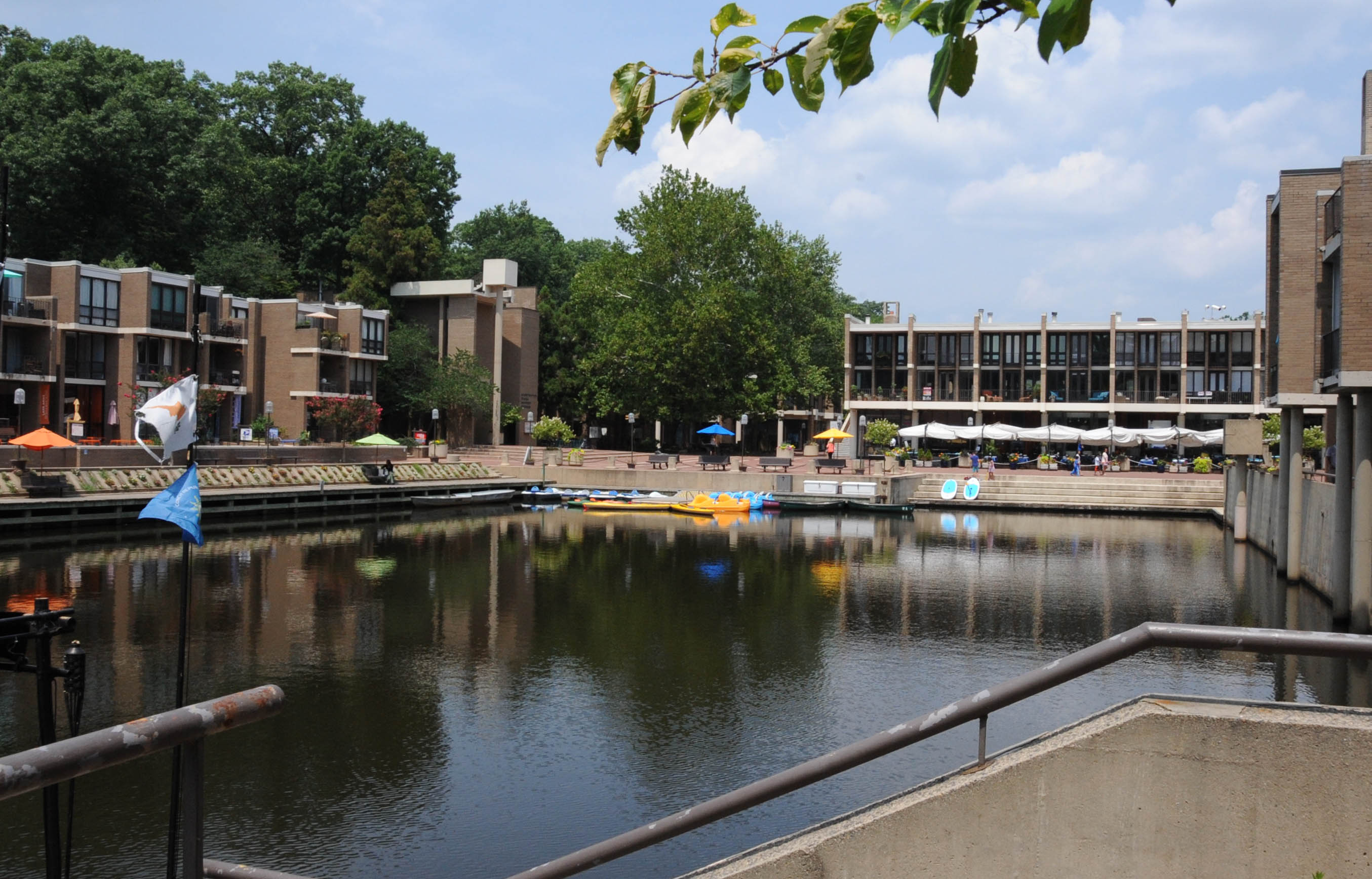 A man-made lake surrounded by brick residential and commercial buildings with paddle boats and umbrellas along the waterfront.