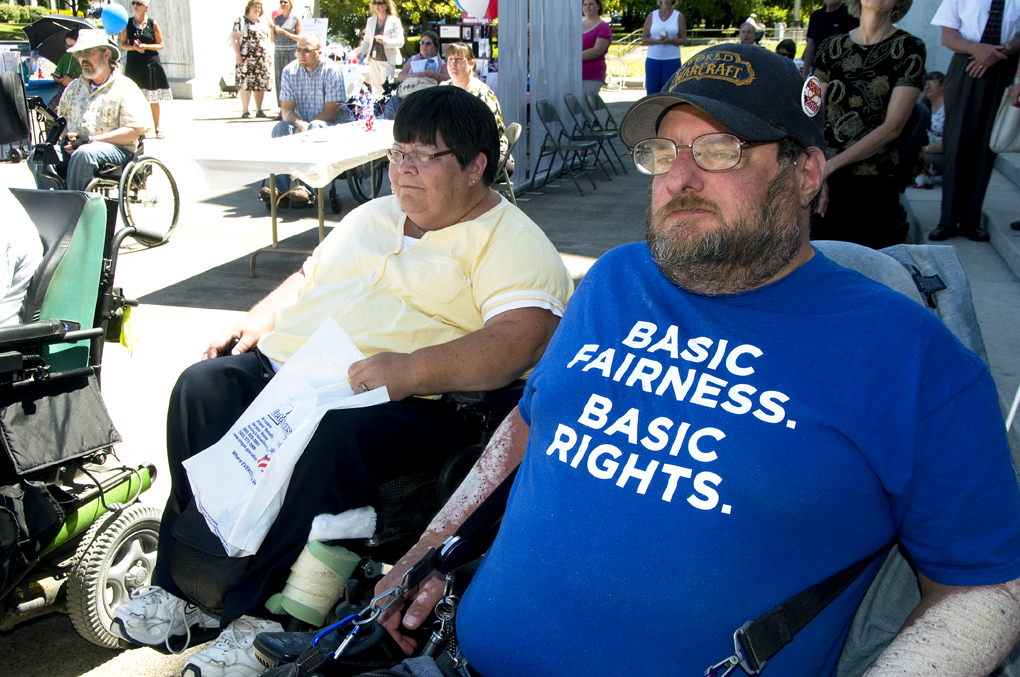 Two people seated outdoors at an ADA event, a power wheelchair visible in the foreground.