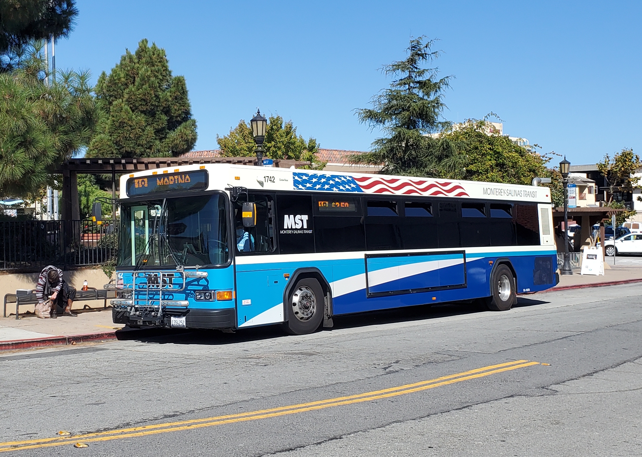 Blue and white MST bus stopped at a transit plaza with trees in the background.