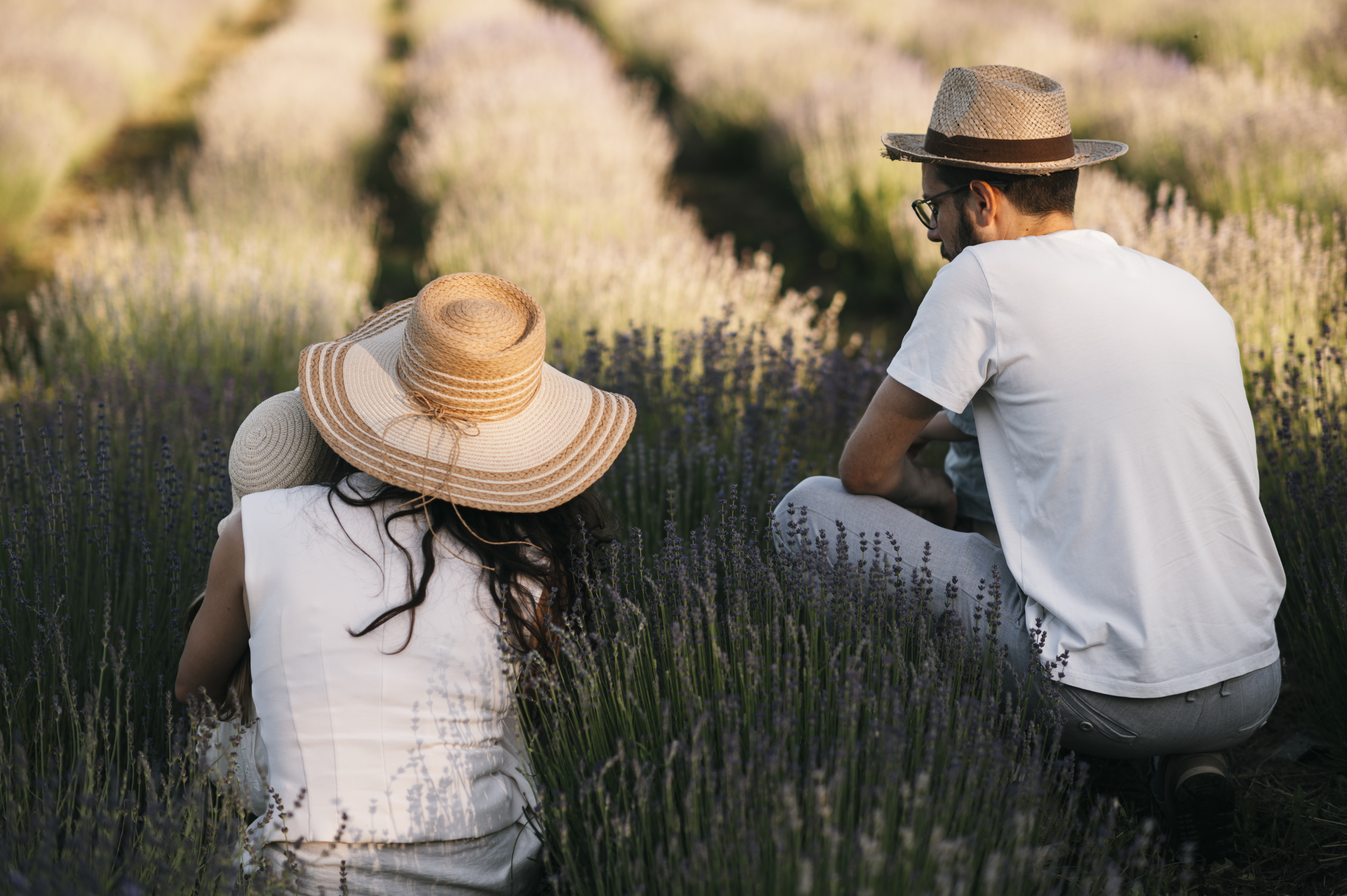 Two people in straw hats sitting among rows of blooming purple lavender in summer sunlight.