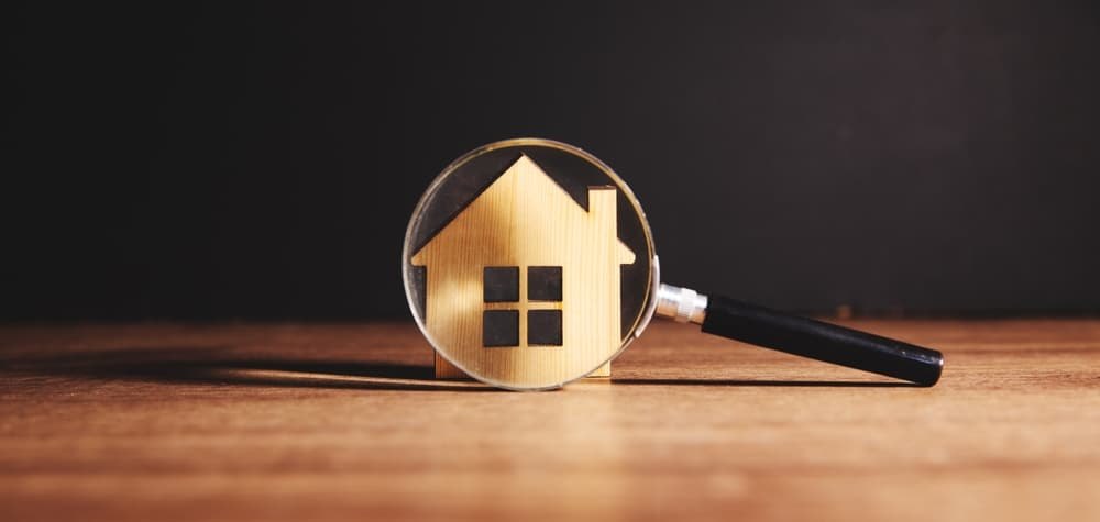Small wooden model house with a magnifying glass in front of it on a wooden surface.