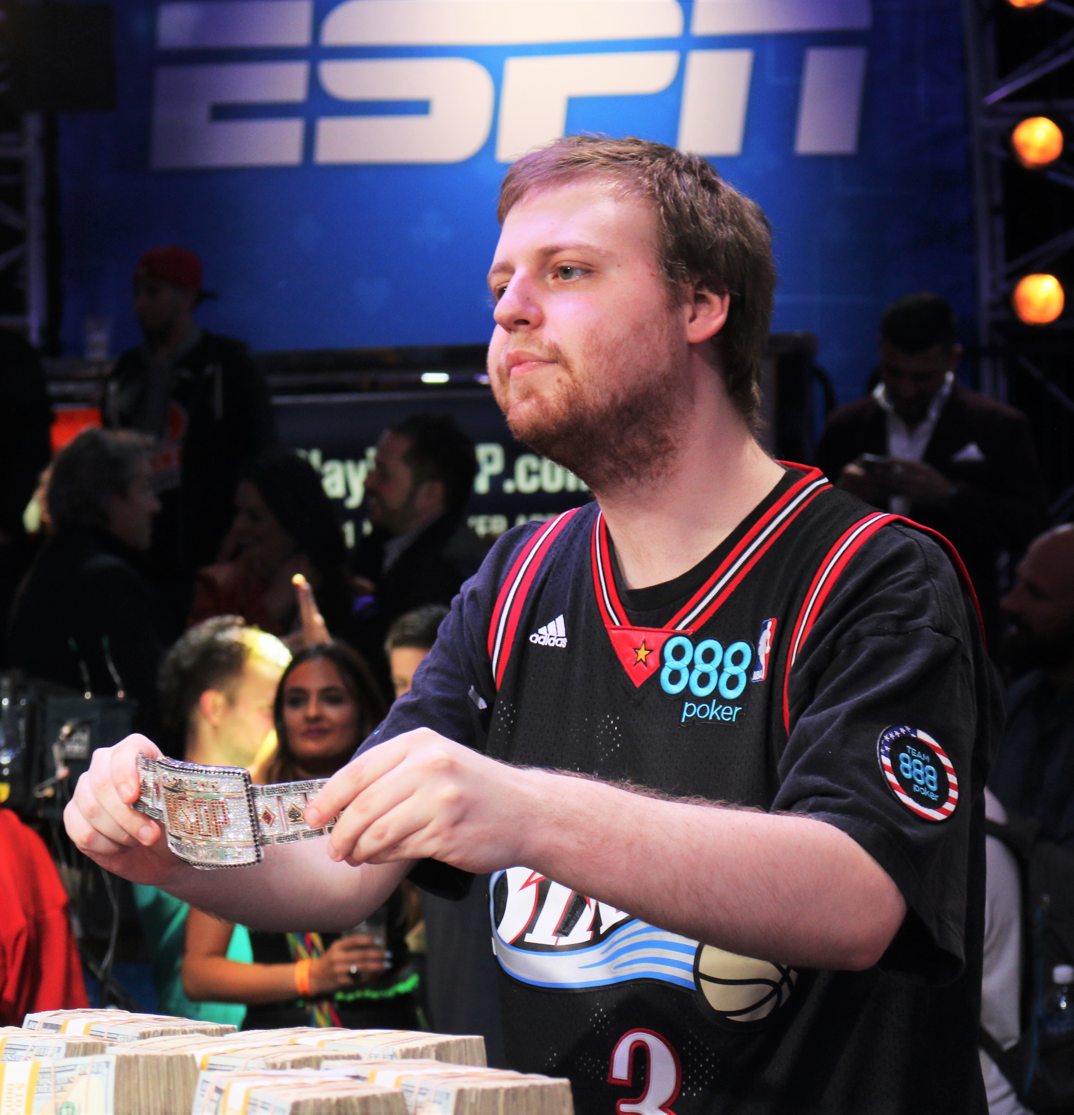 Man in a poker jersey holding cash in front of an ESPN banner at a tournament.