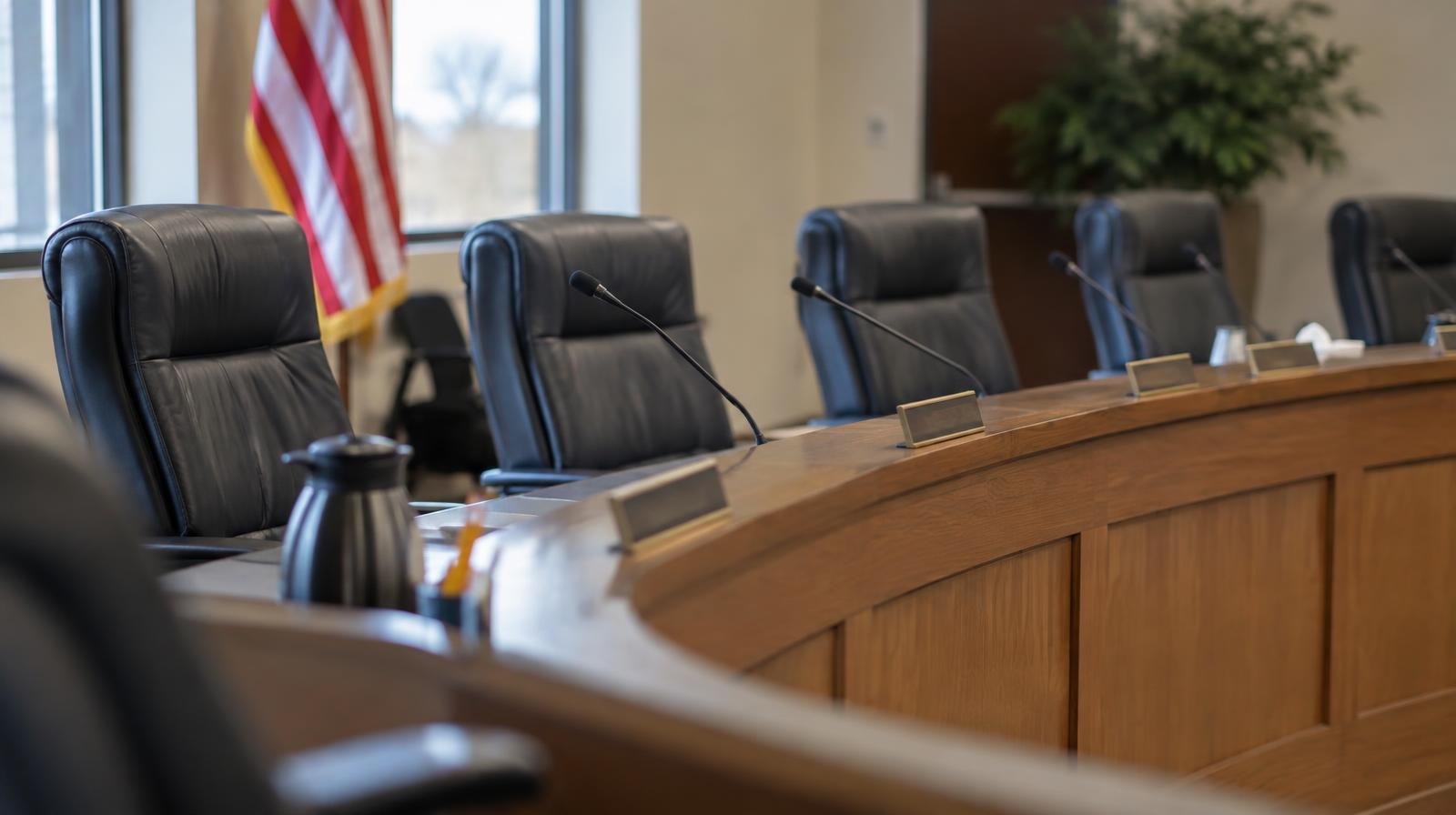 Empty chairs at a curved school board meeting dais with name placards and microphones.