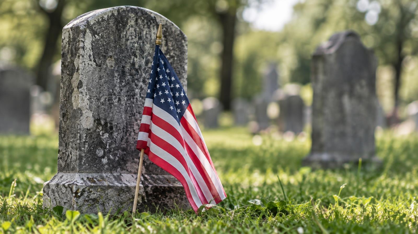 Small American flag planted before a weathered granite headstone on green grass.
