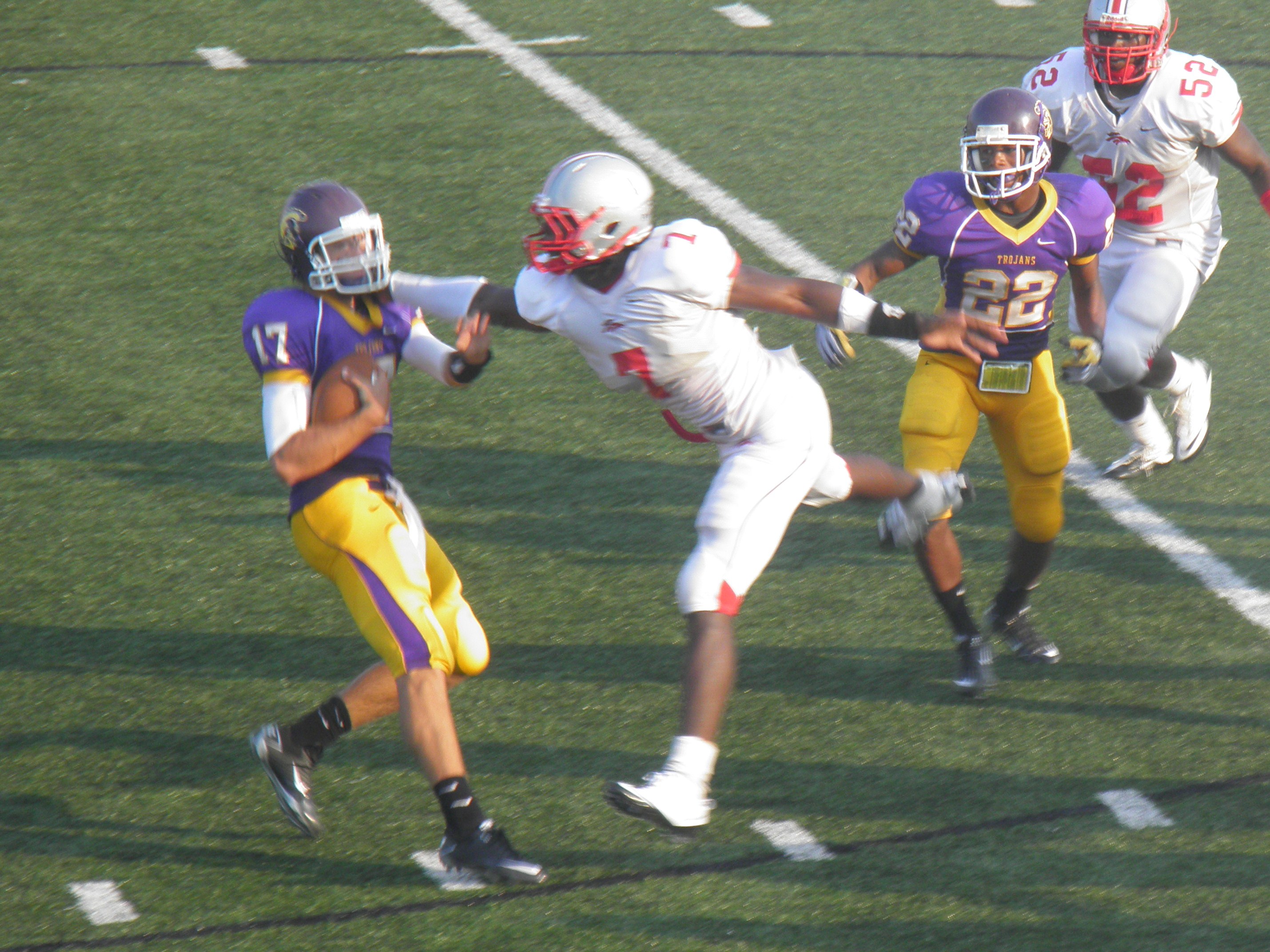 Football players in purple and gold uniforms face off on an artificial turf field.