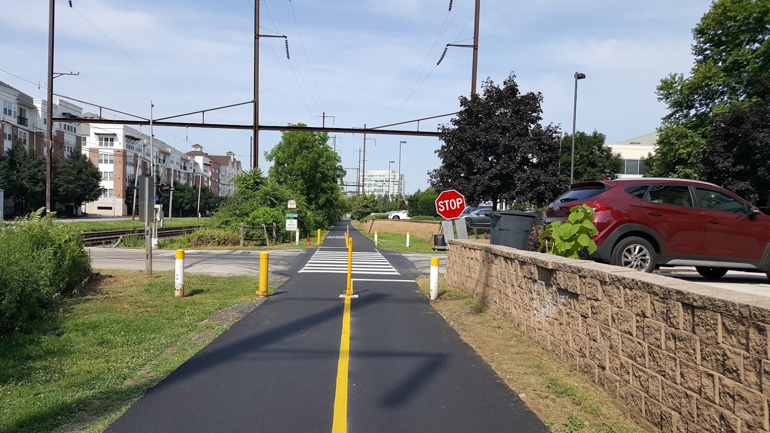 Paved multi-use trail with yellow center stripe and bollards at a suburban road crossing.