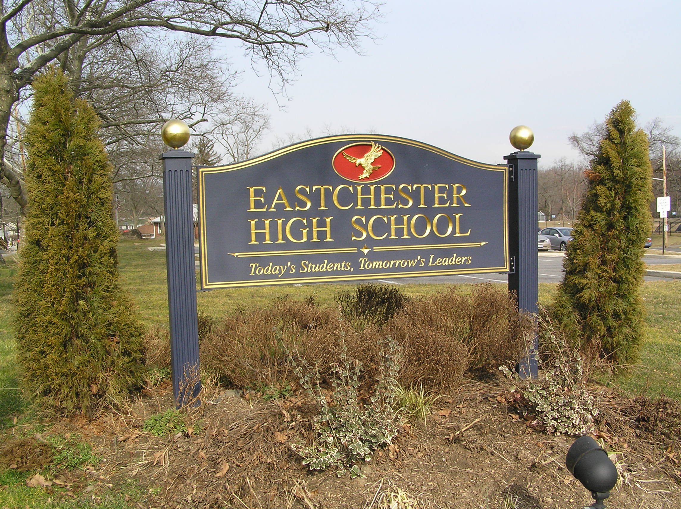 Dark entrance sign reading Eastchester High School flanked by posts with gold finials and evergreen shrubs.