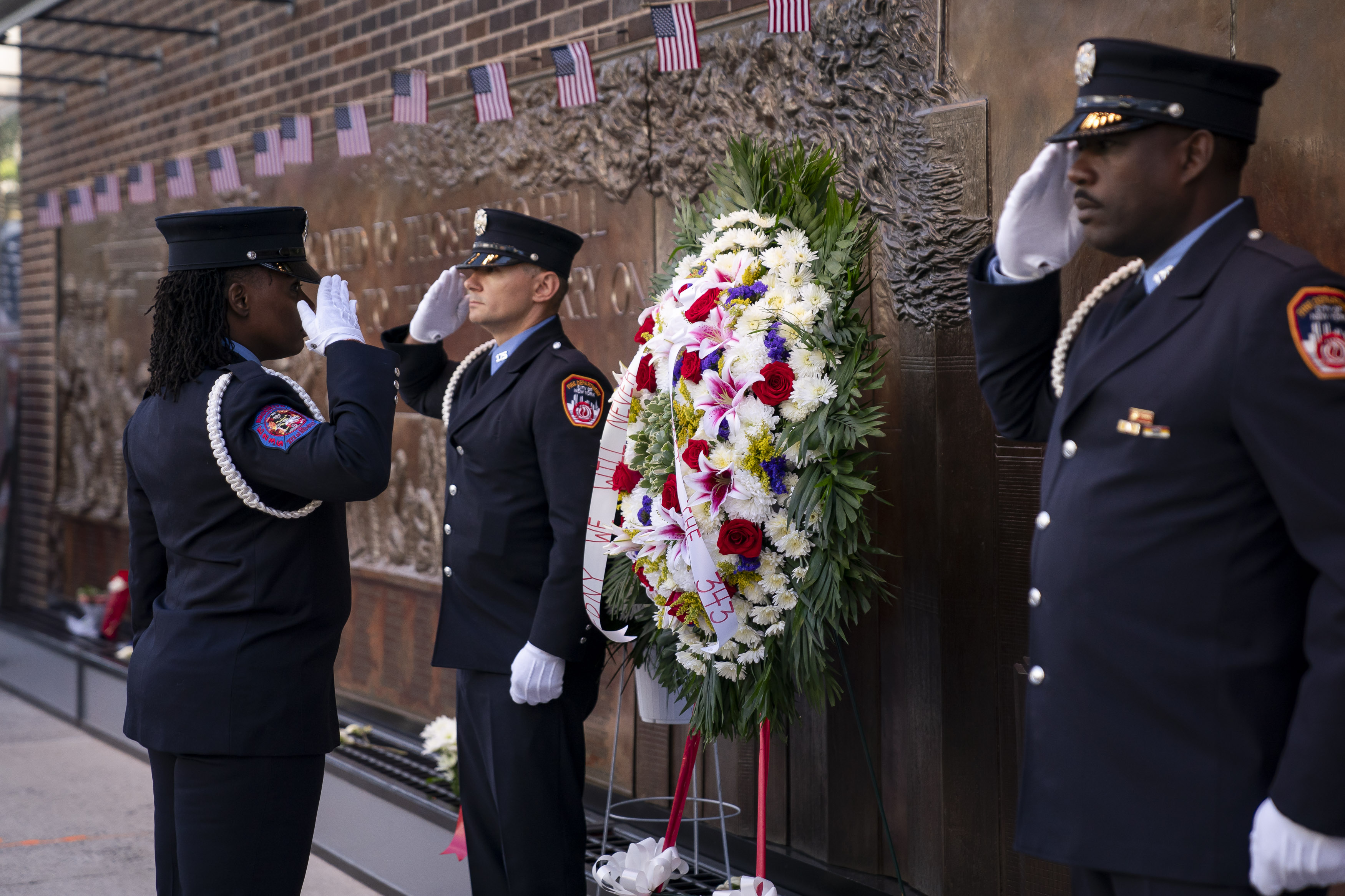 Three firefighters in dress uniforms saluting beside a large floral wreath at a memorial wall.