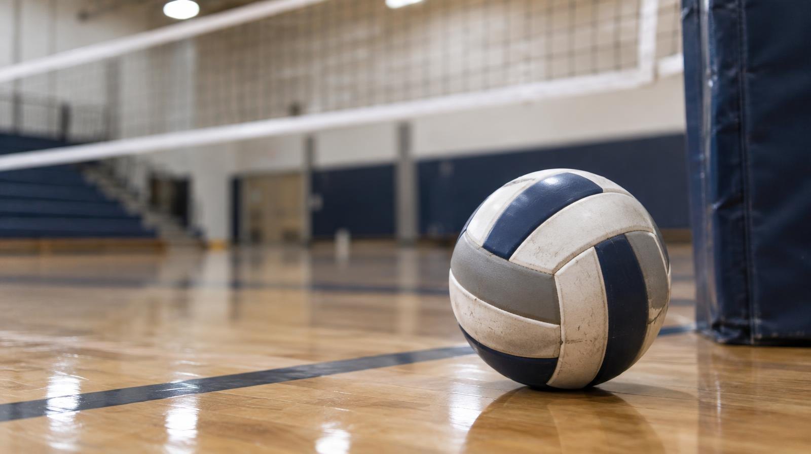 A volleyball resting on a hardwood gymnasium floor near a net post.