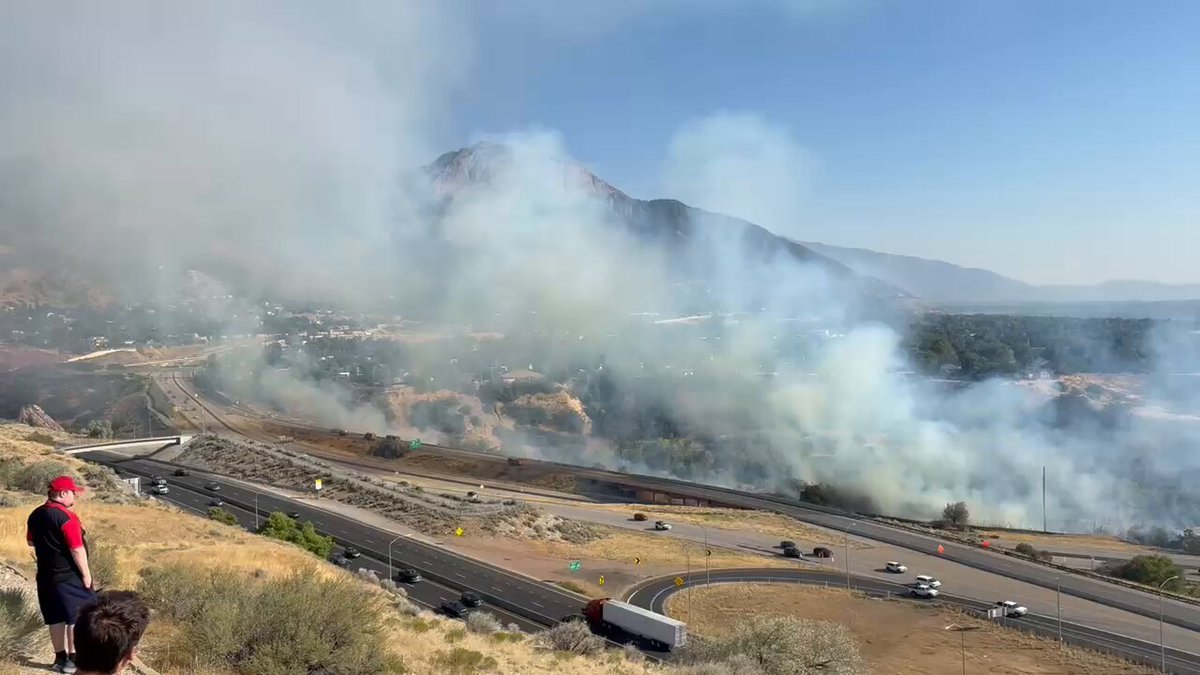 Two people watch thick smoke rise from a wildfire burning along a freeway interchange.