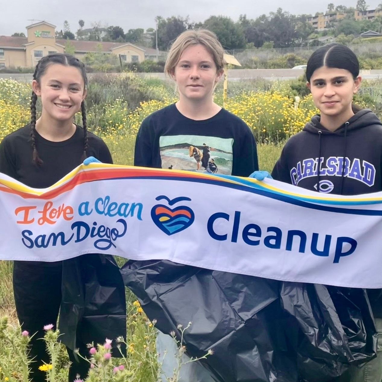 Three young volunteers hold a cleanup event banner outdoors in a field of yellow wildflowers.