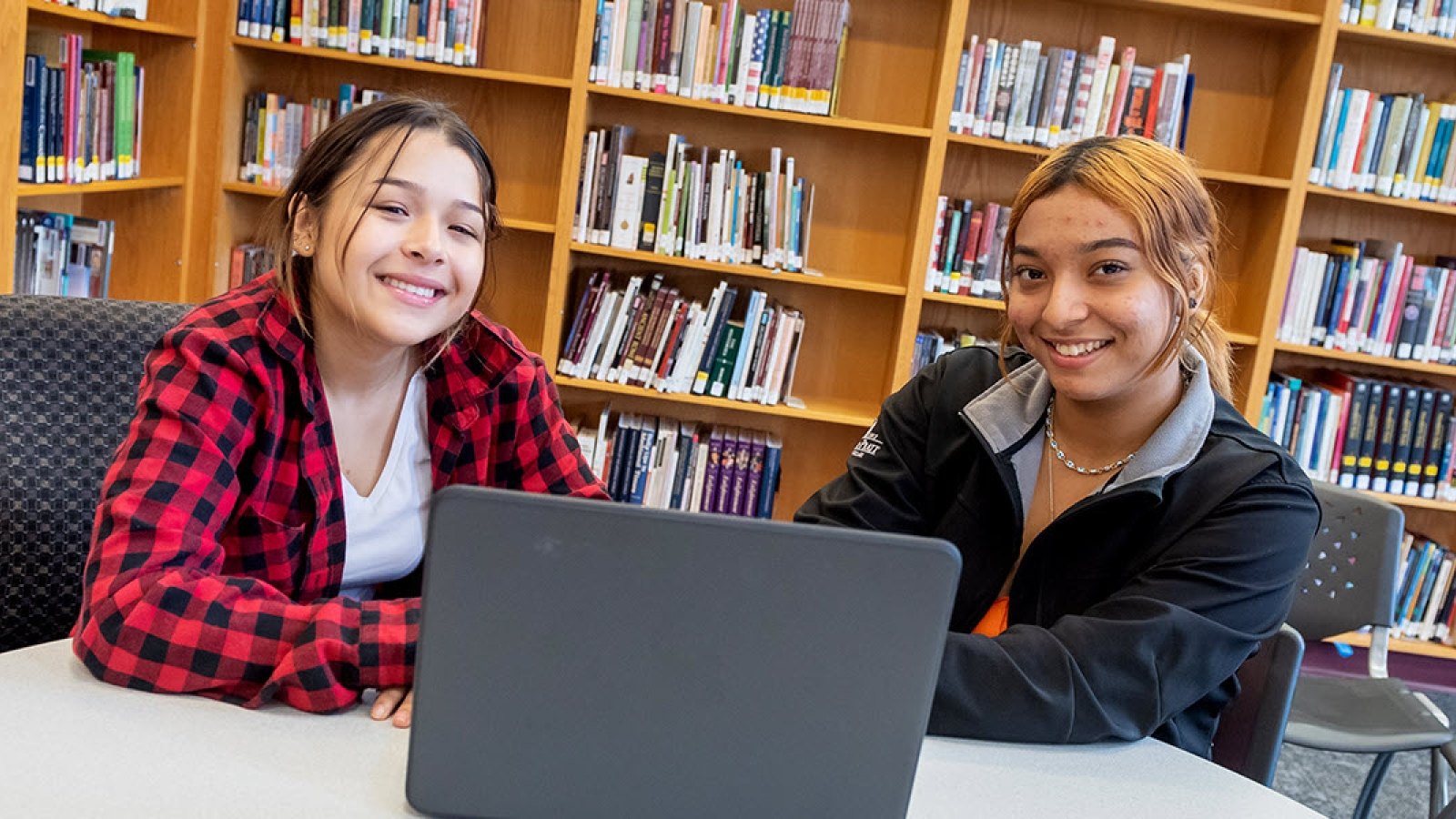 Two smiling teenage girls sit at a library table with an open laptop, bookshelves behind them.