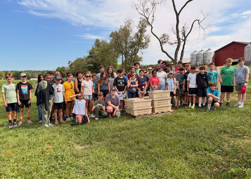 Large group of middle school students posing outdoors on a farm field with wooden crates and a red barn in the background.