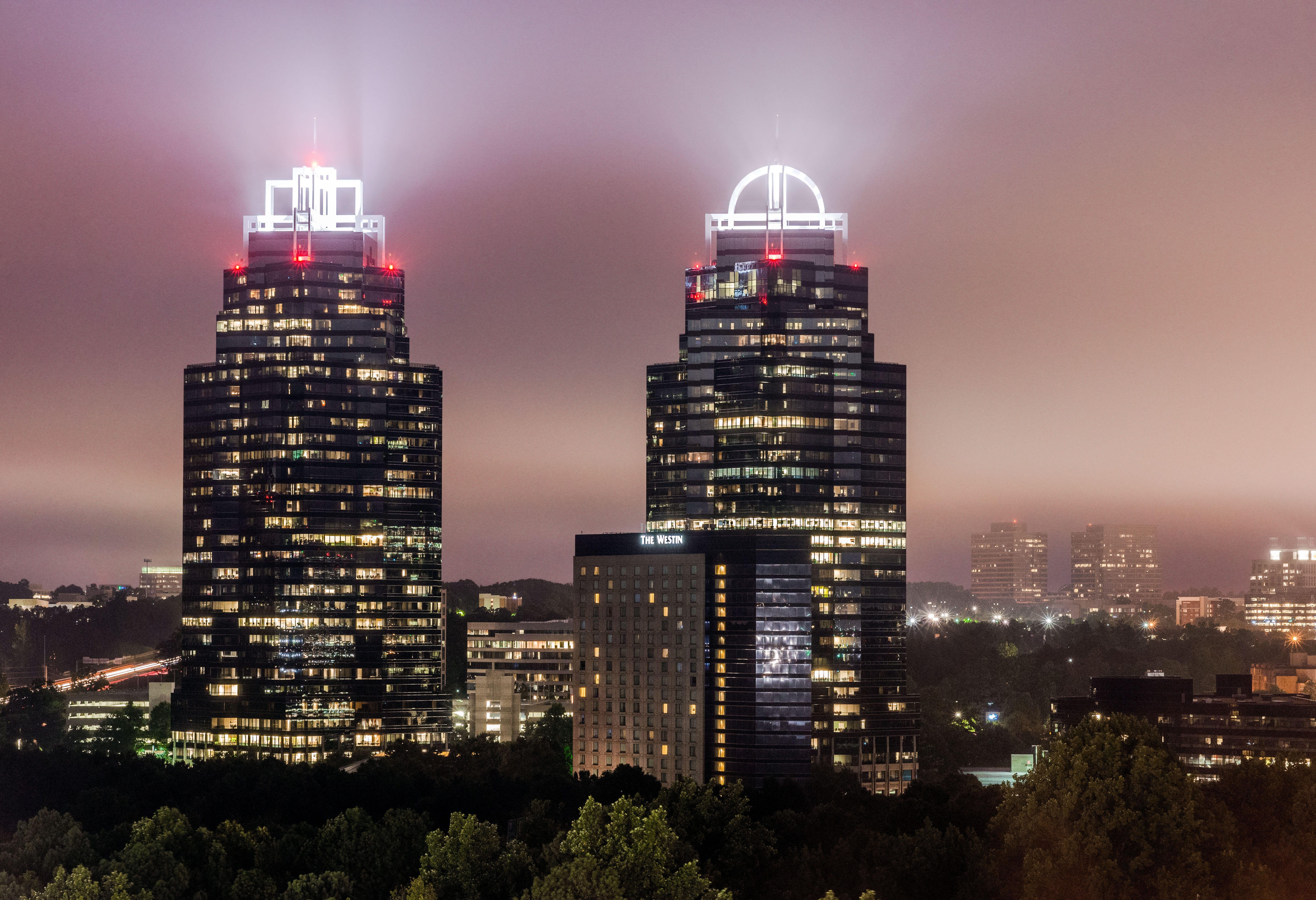 Two illuminated glass skyscrapers at night surrounded by a city skyline and trees.