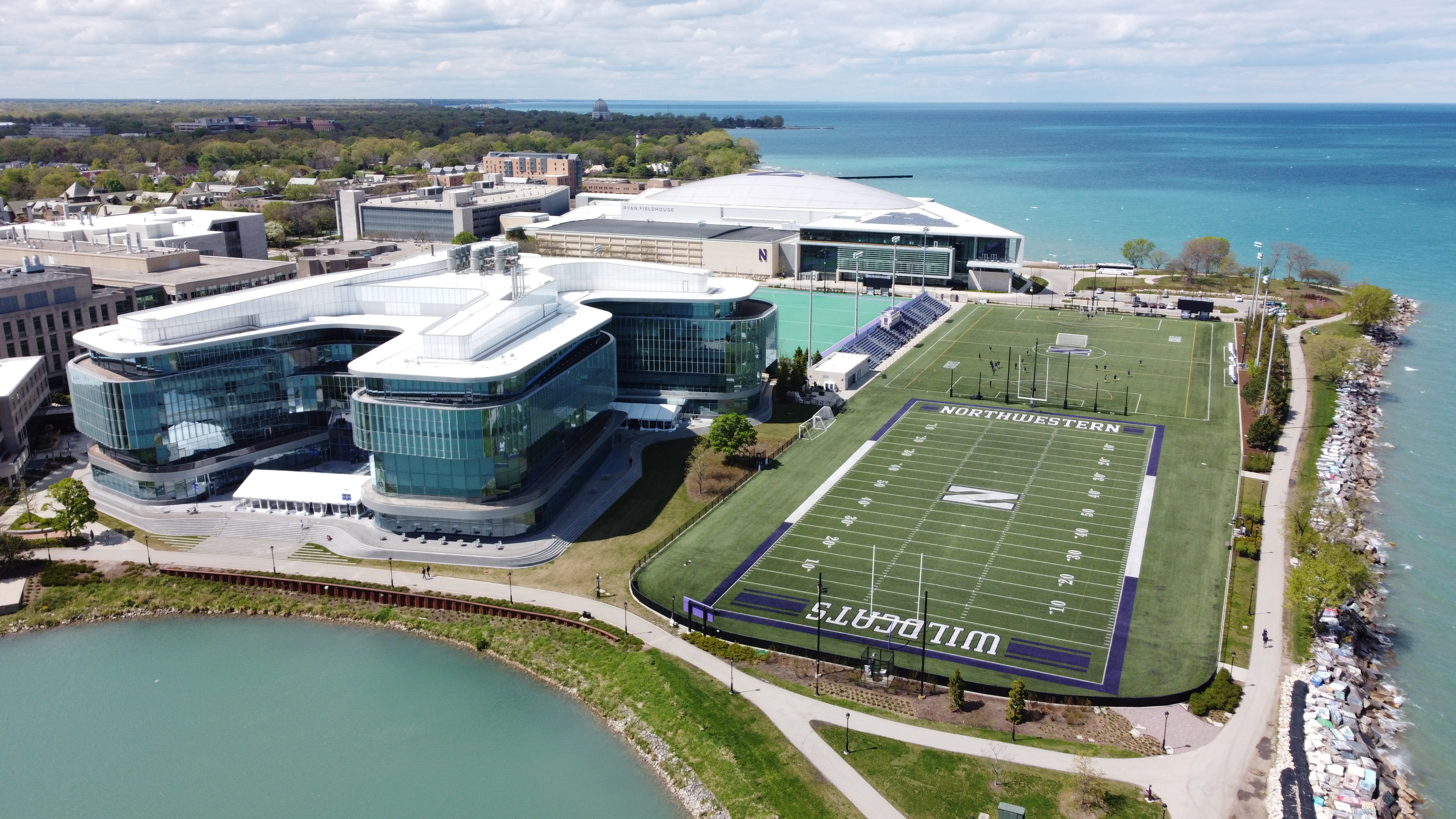 Aerial view of a university athletic campus along a lake shoreline with large modern buildings and practice fields.