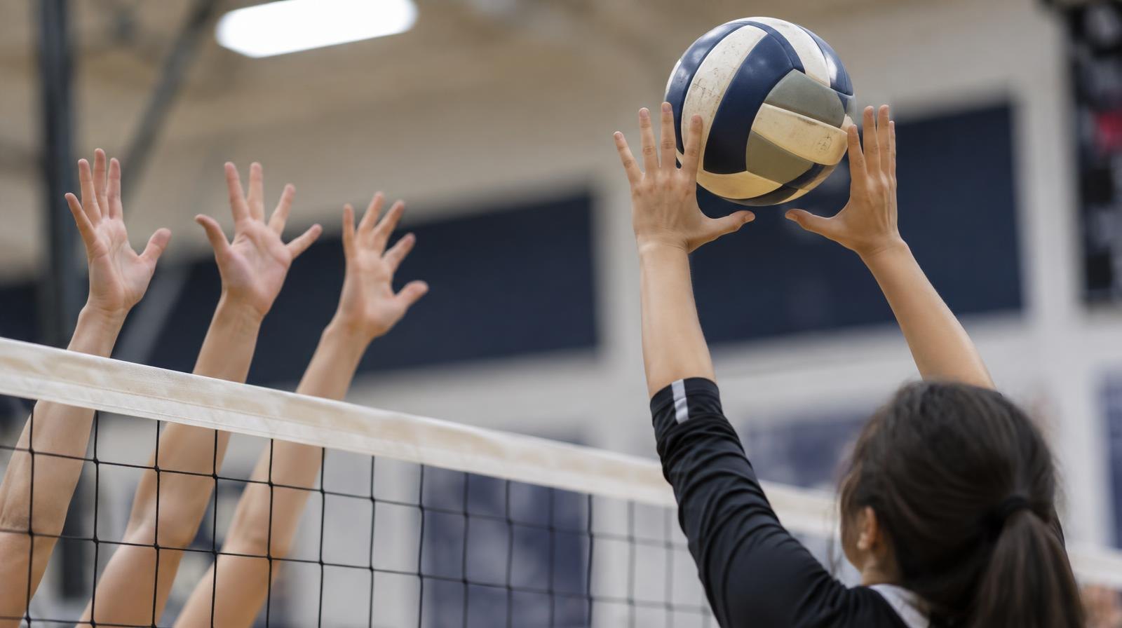 A volleyball above a net with players' hands reaching up to block in a gymnasium.