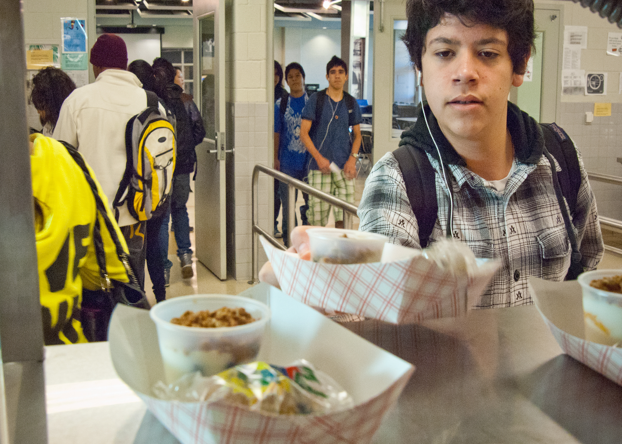 A student in a plaid shirt holds a food tray in a school cafeteria serving line.