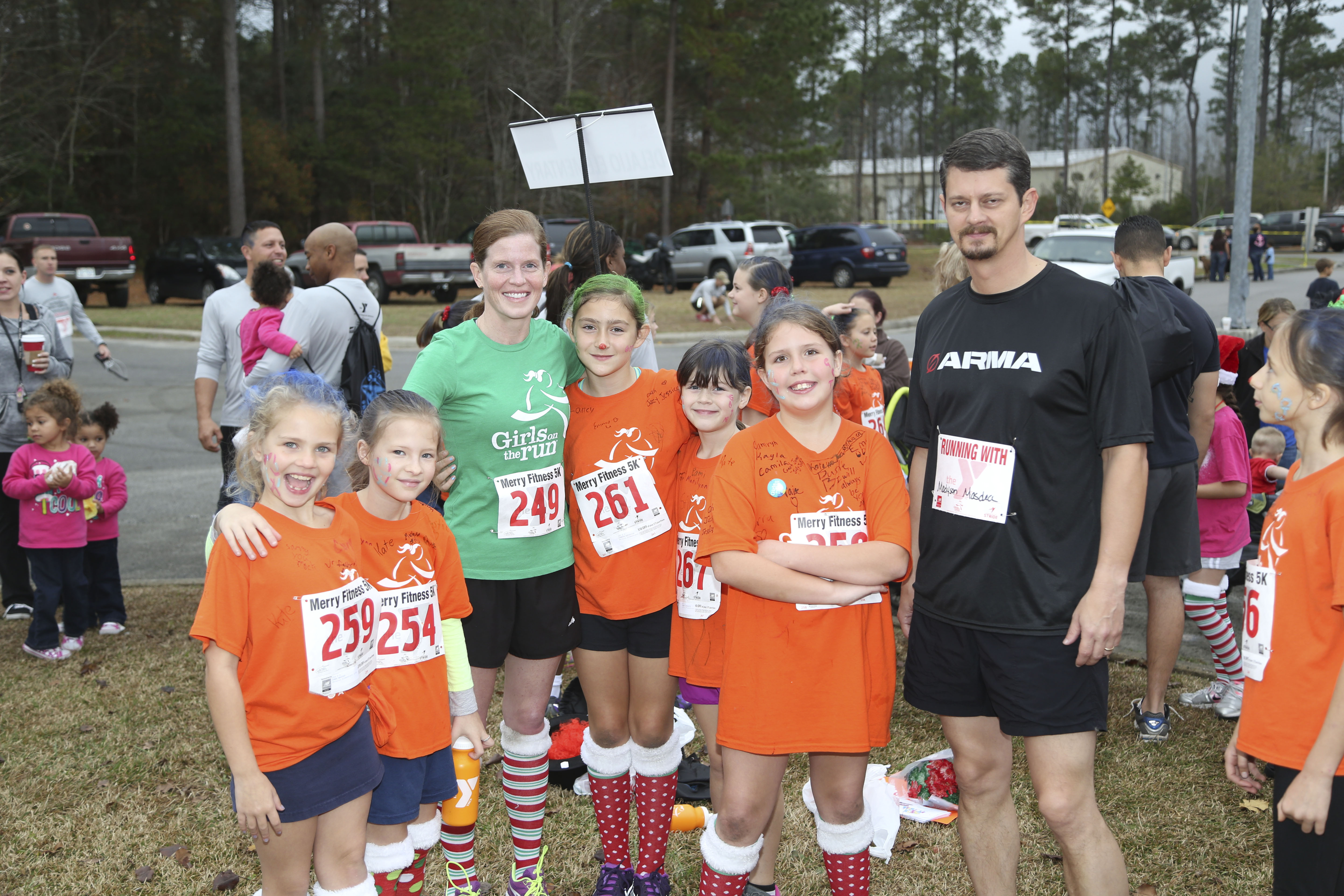 Girls in orange race shirts and numbered bibs pose outdoors with two adults.