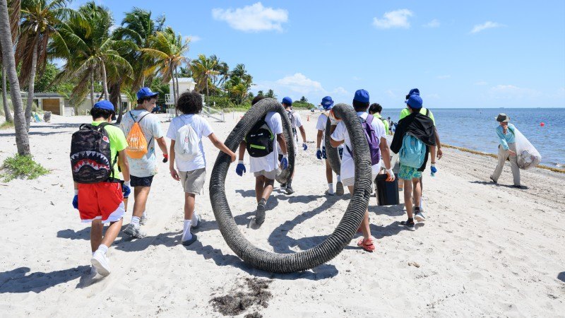 Volunteers in blue caps carry a large coiled hose along a sandy beach near palm trees.