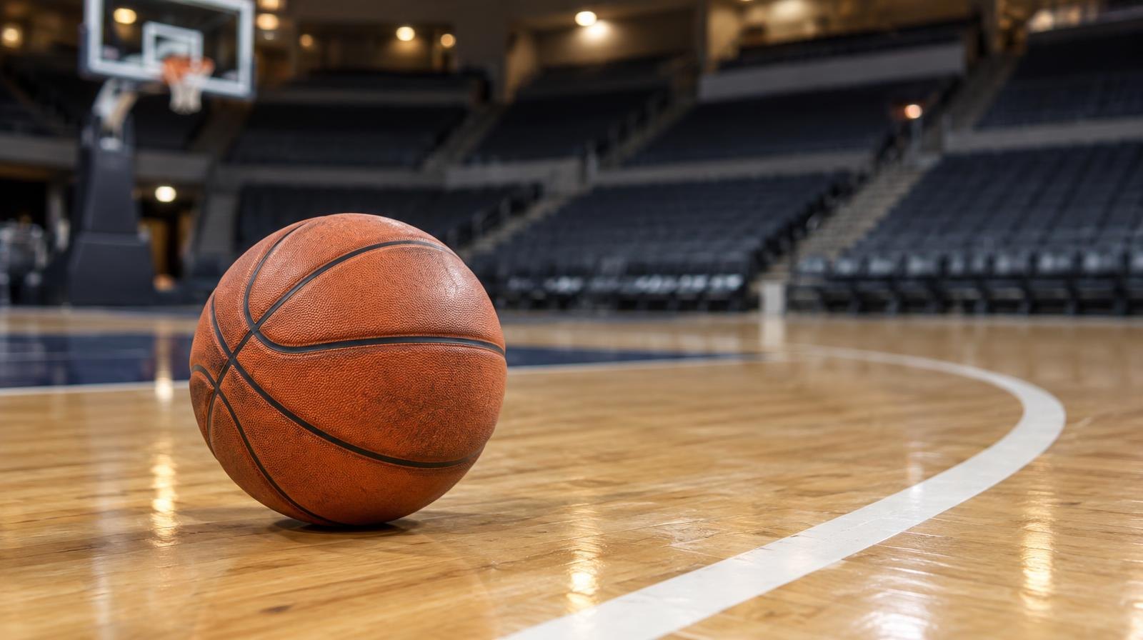 A basketball resting on a hardwood court near the three-point arc with empty arena seats behind it.