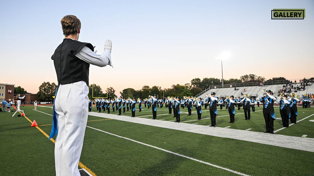 Drum major leads a marching band in blue and white uniforms on a football field at dusk under stadium lights.