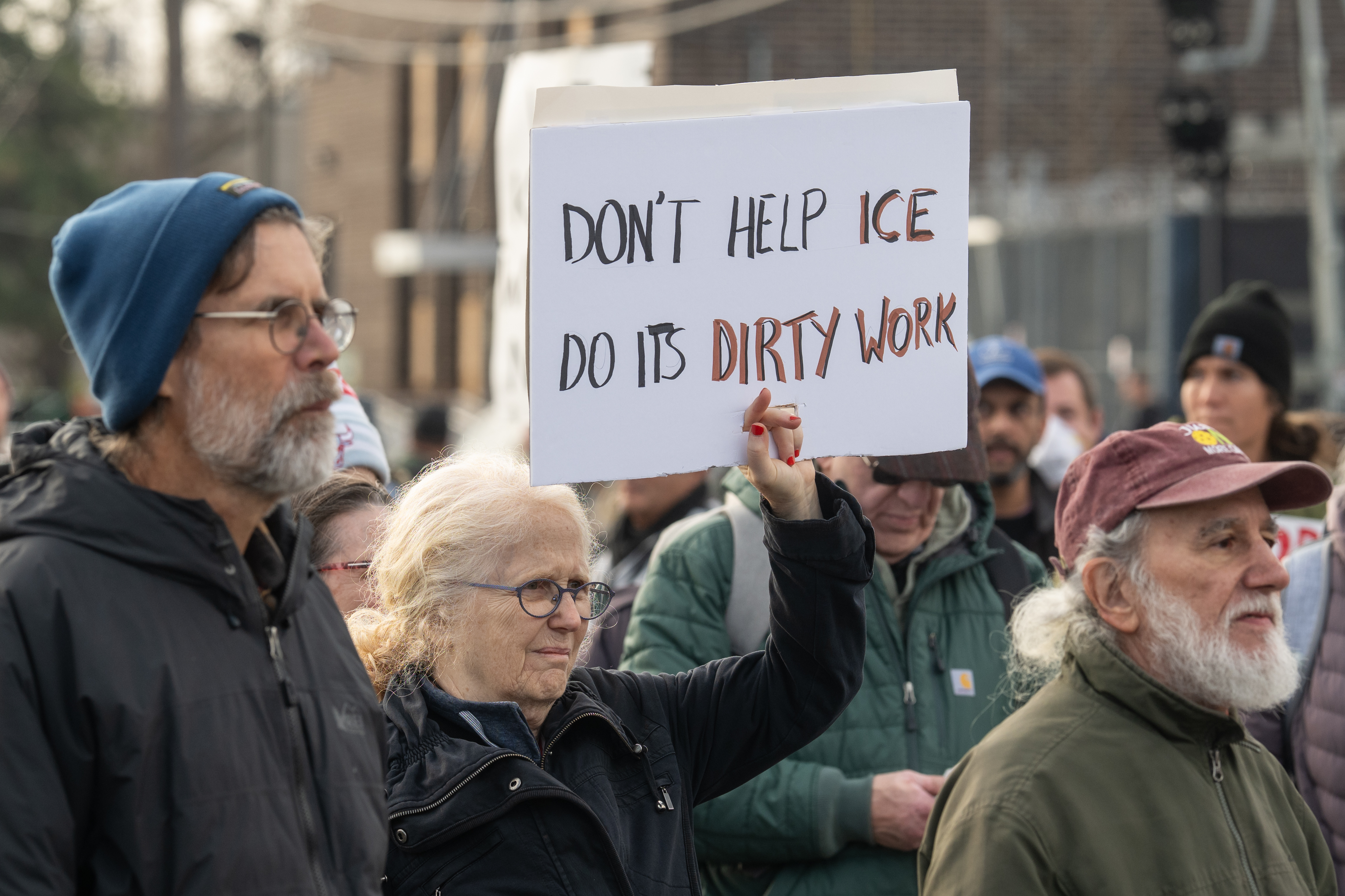 Woman holds a sign reading 'Don't Help ICE Do Its Dirty Work' at an outdoor protest.